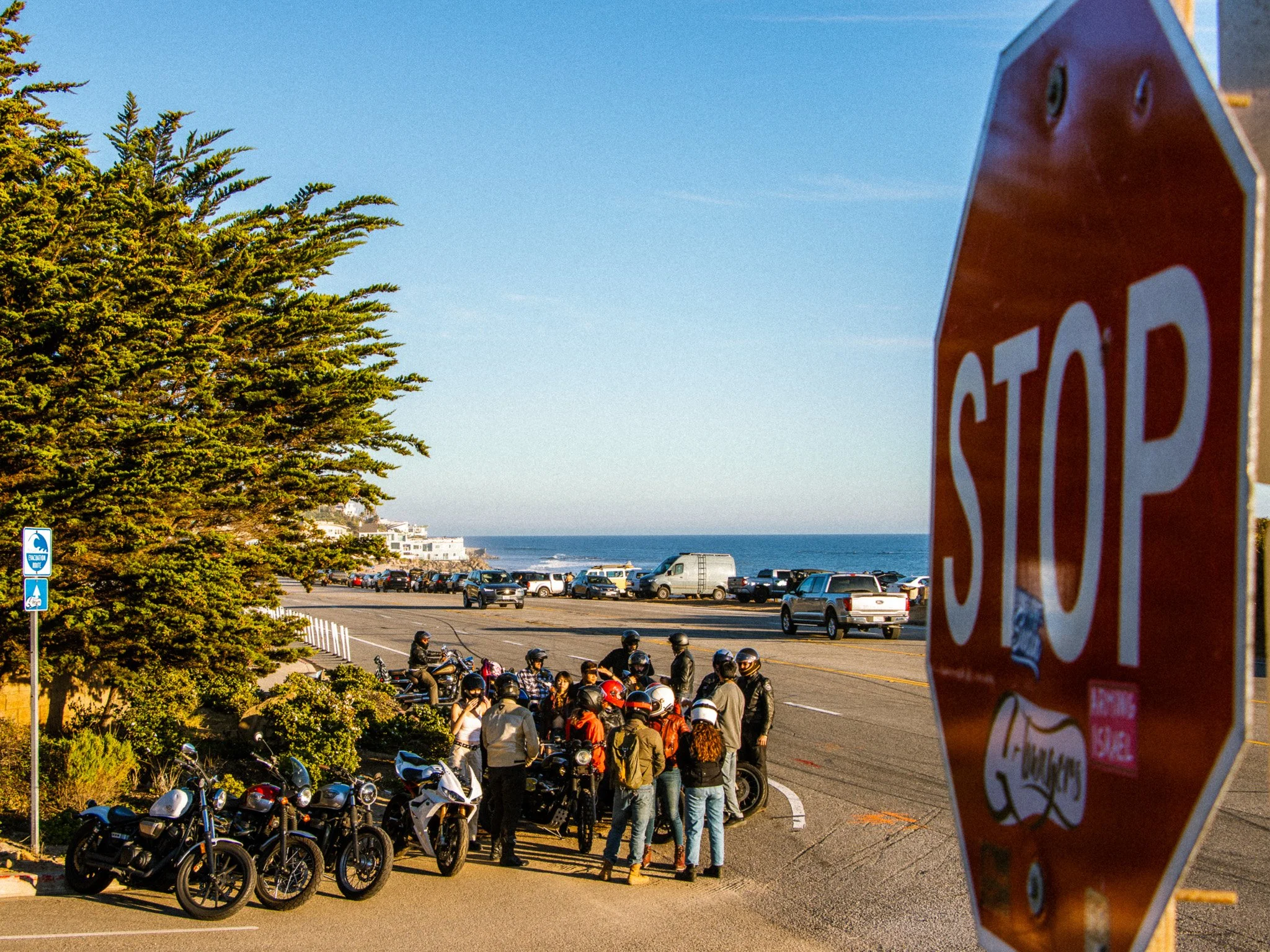 Crowd of people and motorbikes near a cliffside, with the ocean in the background, and a stop sign partially visible on the right.