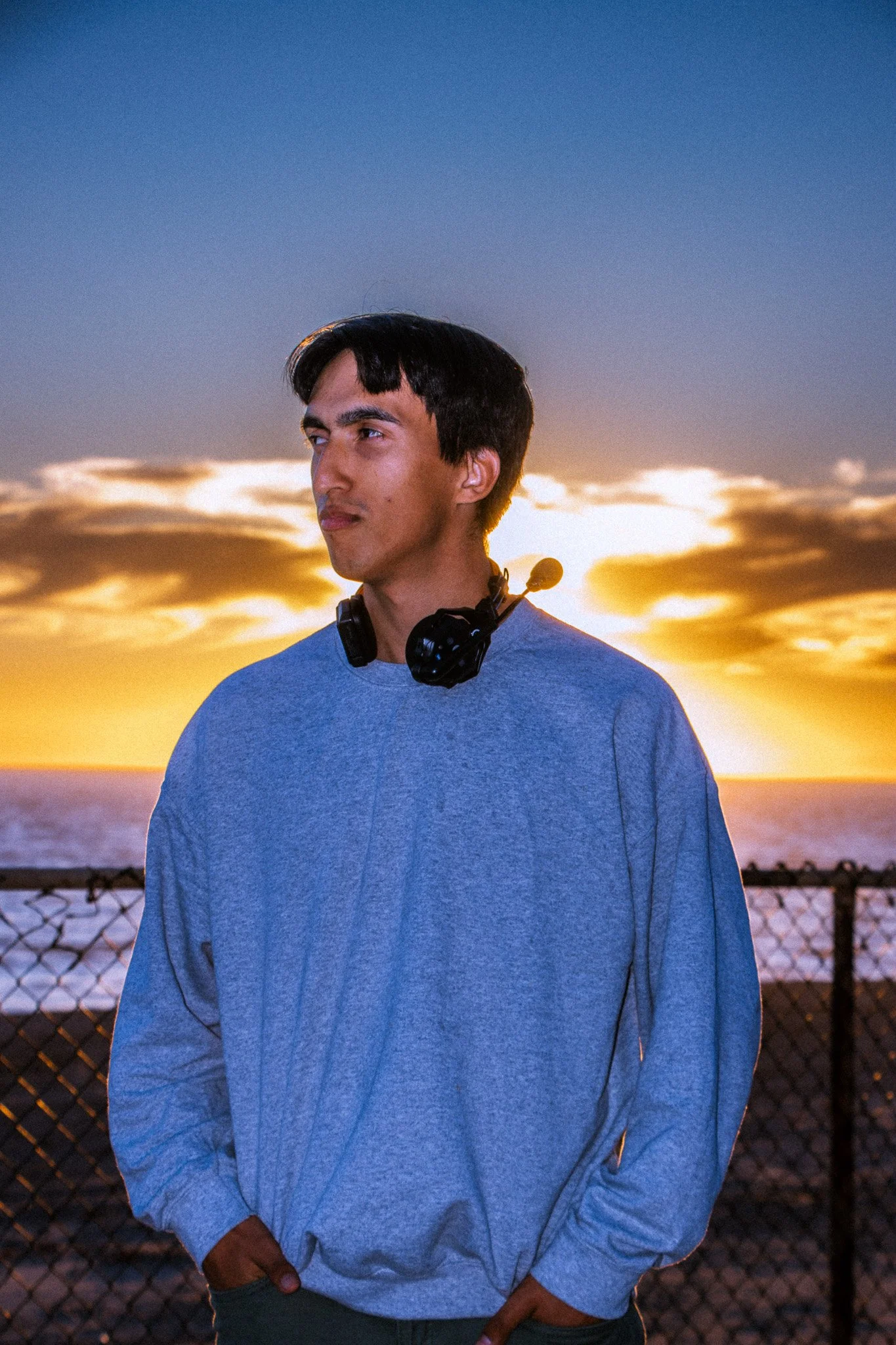 A young man wearing a gray long-sleeve shirt and headphones around his neck standing outdoors at sunset with a fence and ocean in the background.
