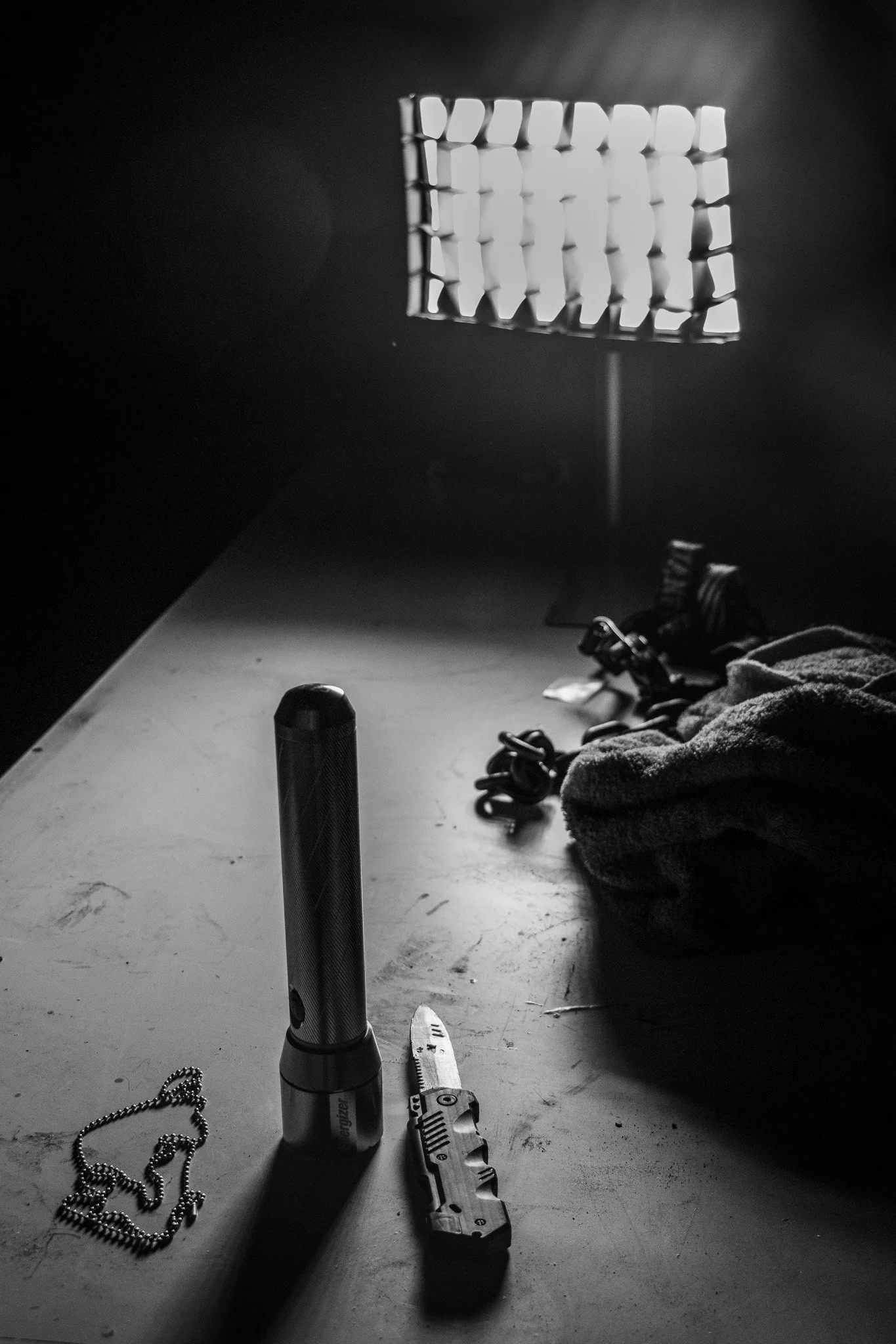 Black and white photo of a desk with various objects, including a flashlight, a small gun, a chain, a towel, chain links, and a knife, with a grid-like light pattern on the wall in the background.