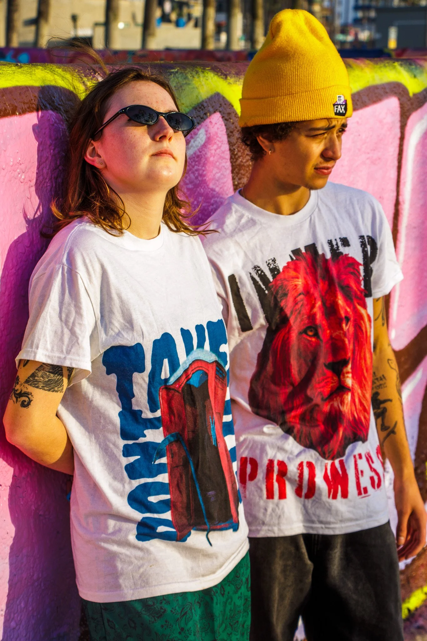 Two young adults standing against a colorful graffiti wall. The girl on the left is wearing sunglasses and a white shirt with a graphic, while the boy on the right is wearing a yellow beanie and a white t-shirt with a lion graphic.