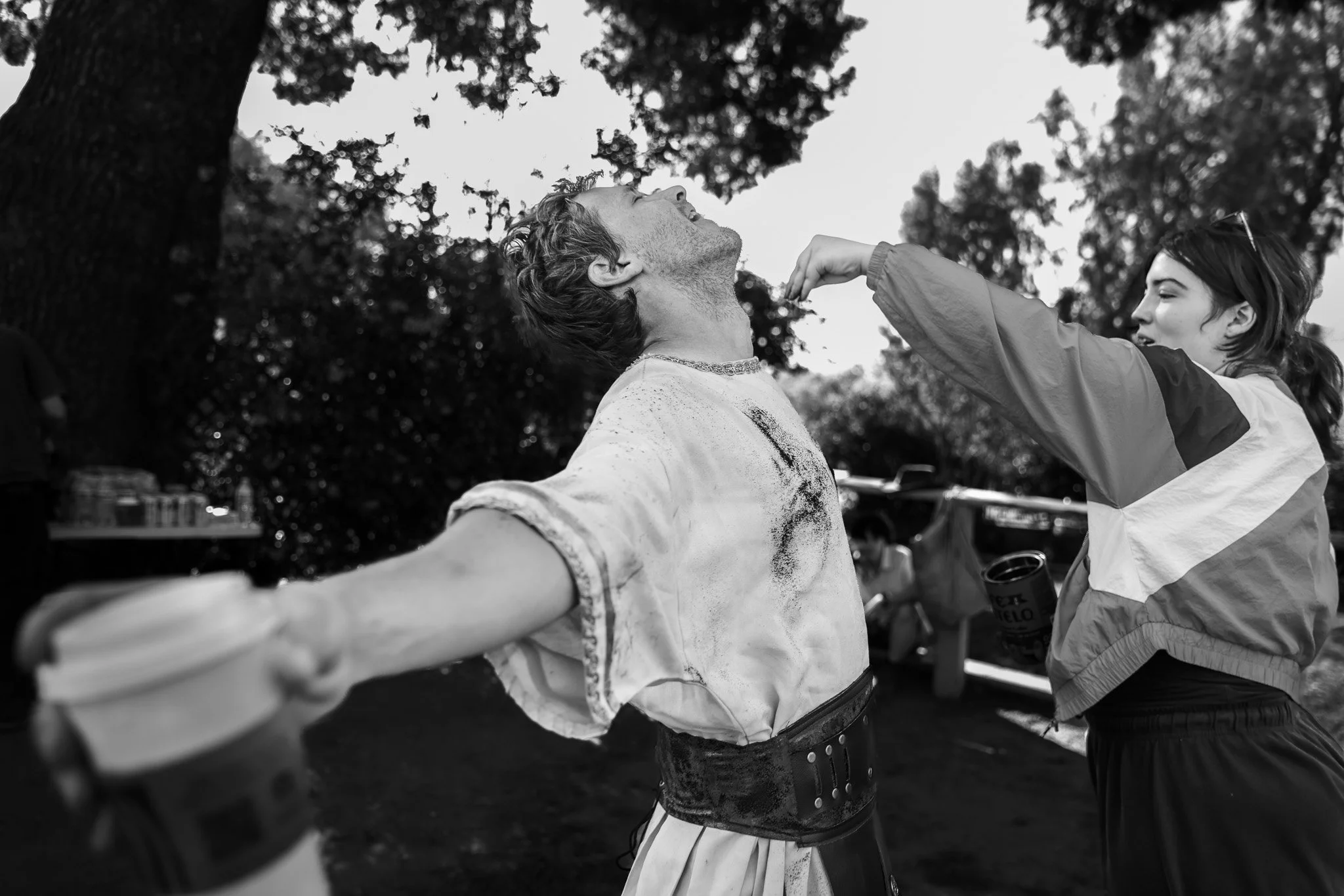 A black-and-white photo of a young man and young woman outdoors surrounded by trees, with the man smiling and the woman reaching out to touch his face.