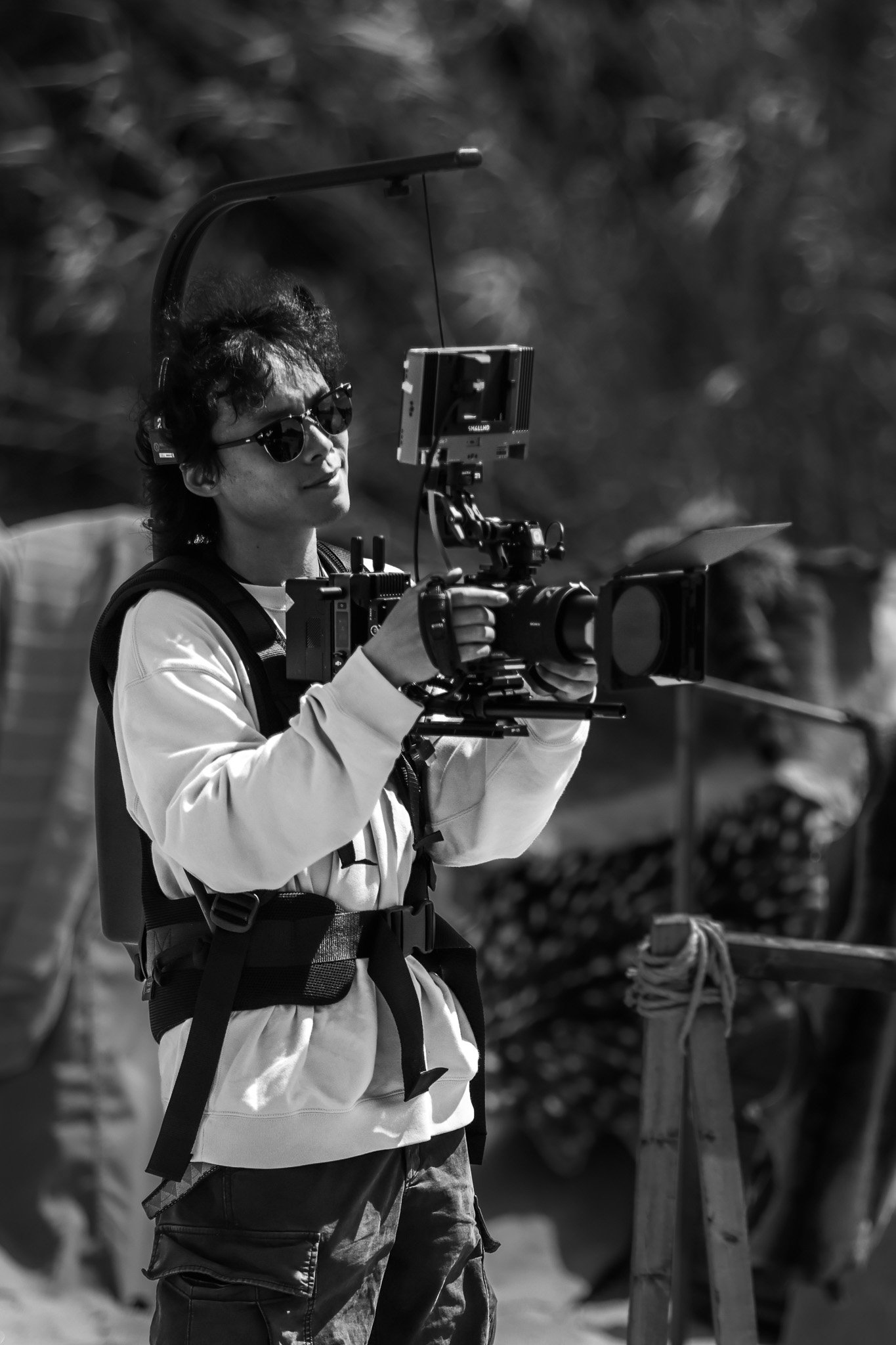 A person with curly hair wearing sunglasses, a white jacket, and a harness, operating a professional video camera outdoors.