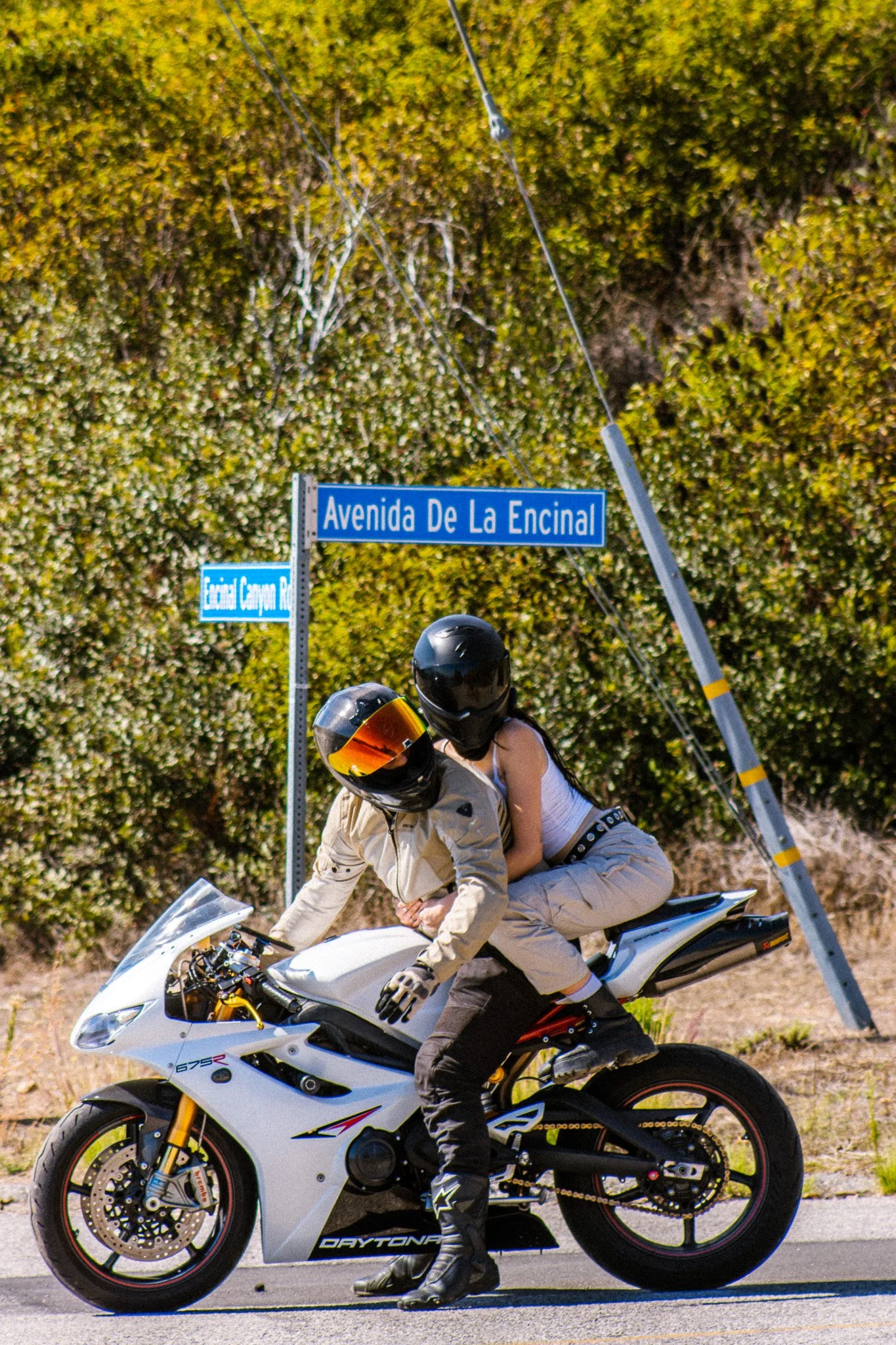 A person in motorcycle racing gear, including gloves and boots, riding a white sports motorcycle with skeletal frame exposed, on a paved road with a hedge and street signs in the background. The street signs read 'Avenida De La Encinal' and 'Ecuador 