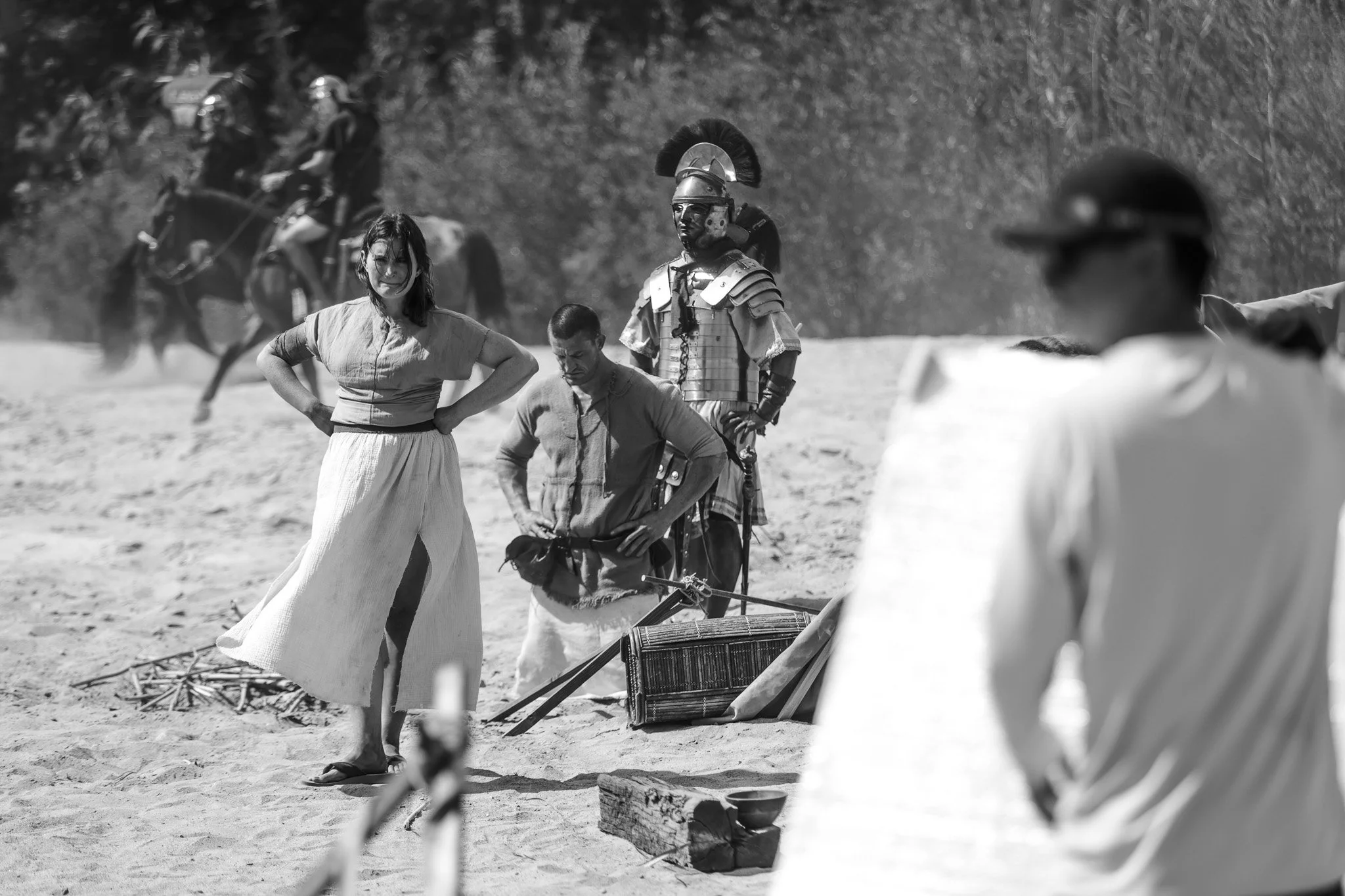 Black and white photo of a woman in a long skirt and blouse, standing with her hands on her hips, on a sandy outdoor set with people and a person in Roman-style armor in the background.