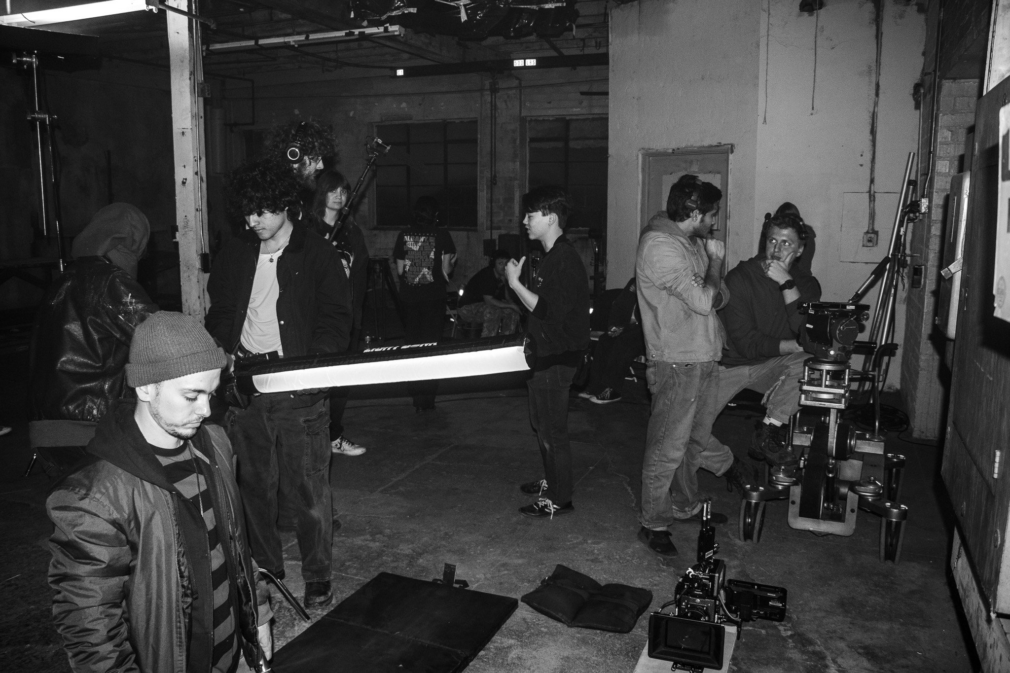A film or photography crew working in a warehouse-like space with equipment including a camera on a dolly and lighting. Several people are engaged in different tasks, some talking or looking at equipment.