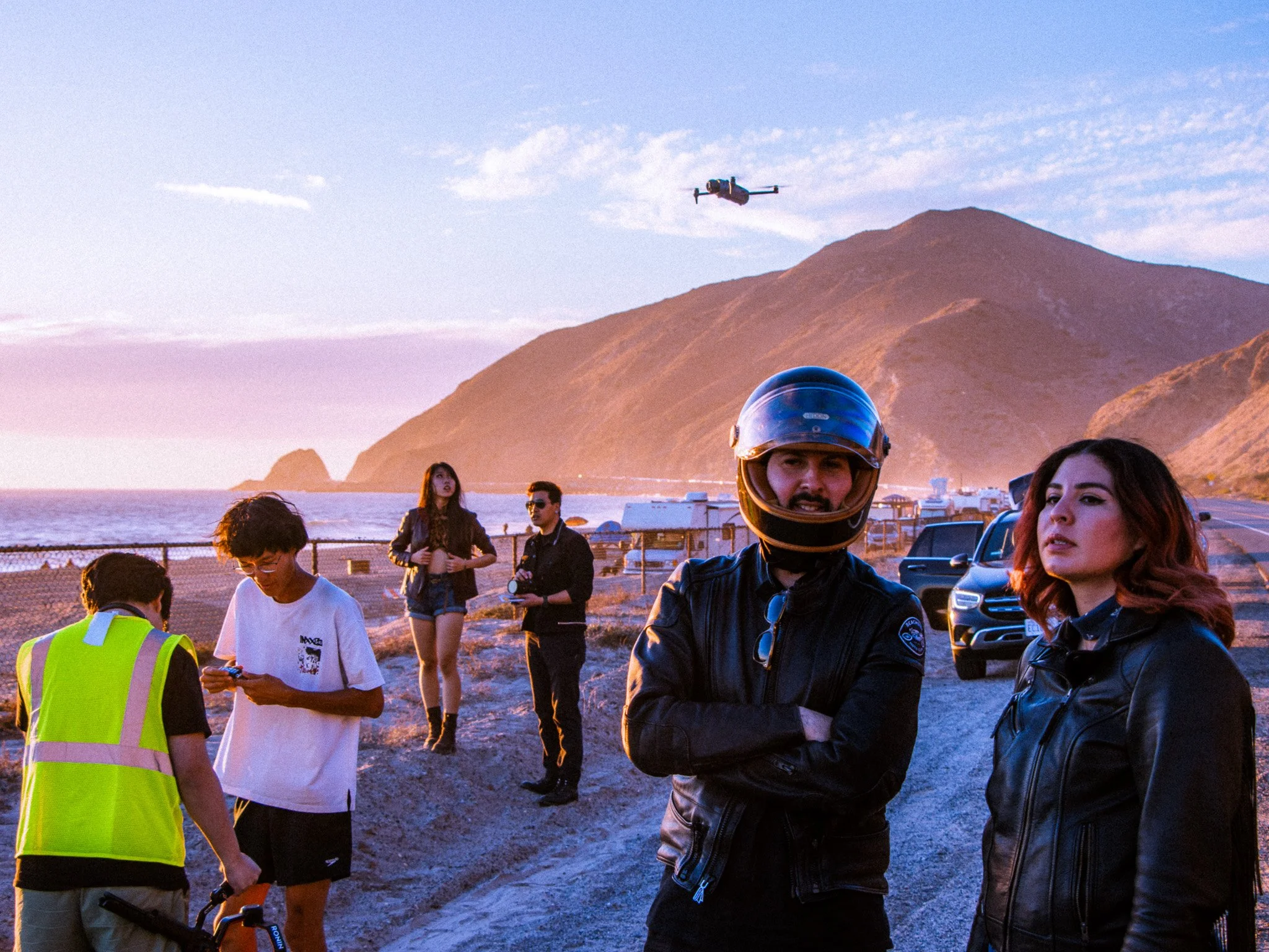 A group of people including a man in a motorcycle helmet and jacket, and others in casual clothing, are gathered outdoors near the coast with mountains in the background at sunset. A drone flies in the sky above them.