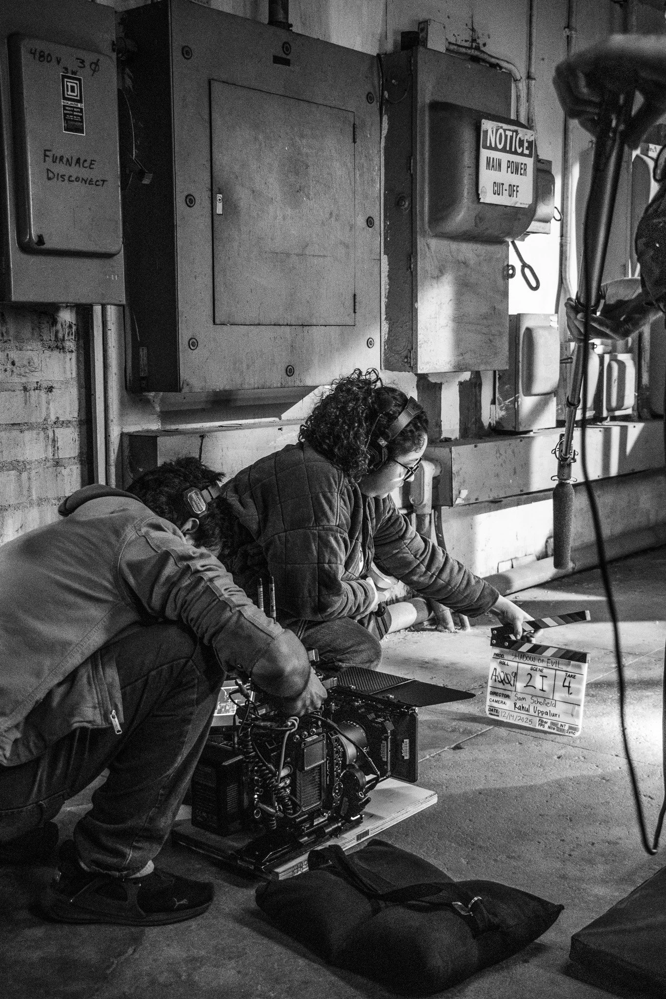 Film crew working on set with camera equipment, clapperboard, and crew members wearing headsets, in an industrial setting with electrical panels and warning signs.