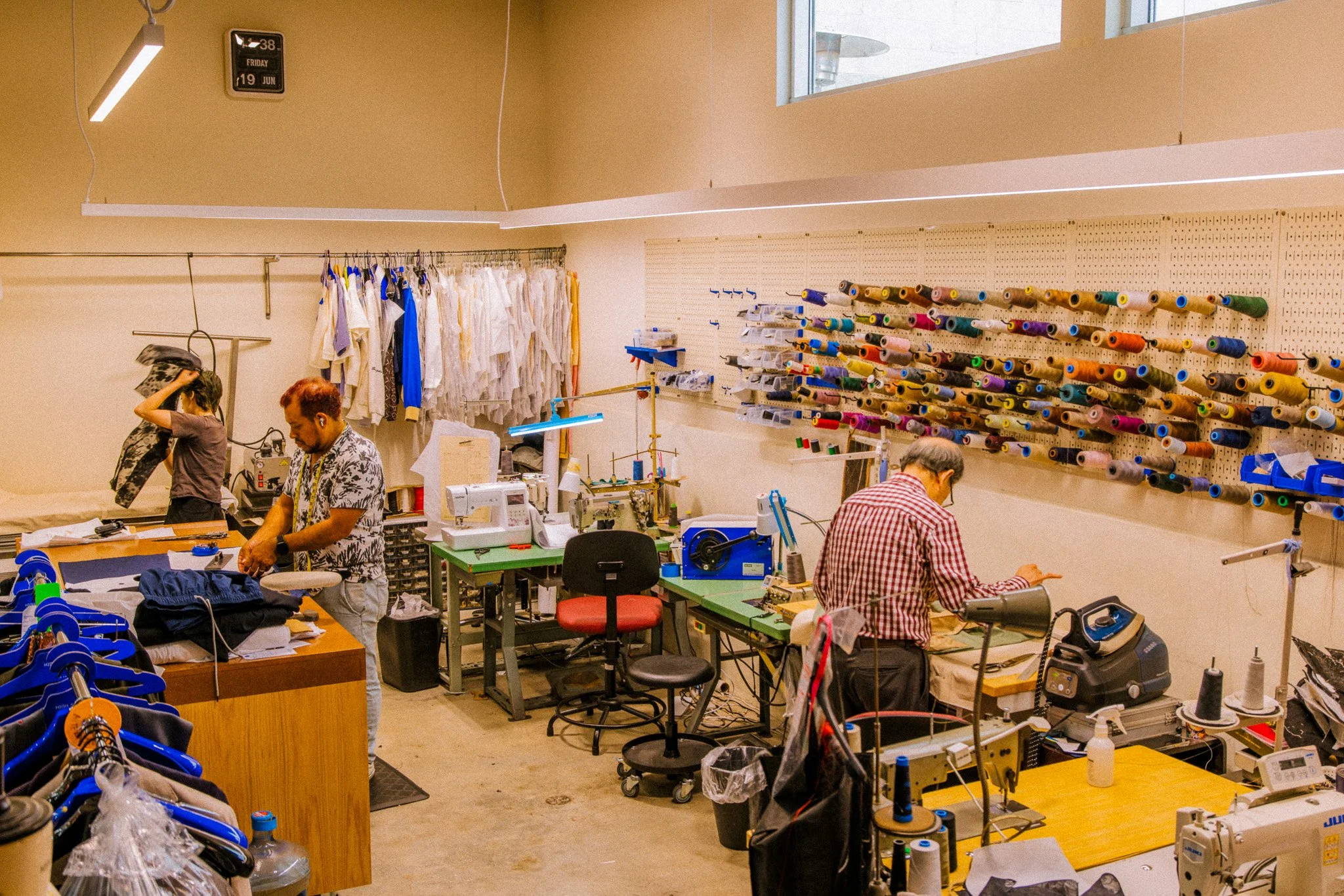 A workspace with sewing and tailoring equipment, including spools of thread on a wall, tables with fabric and sewing machines, and people working on clothing.