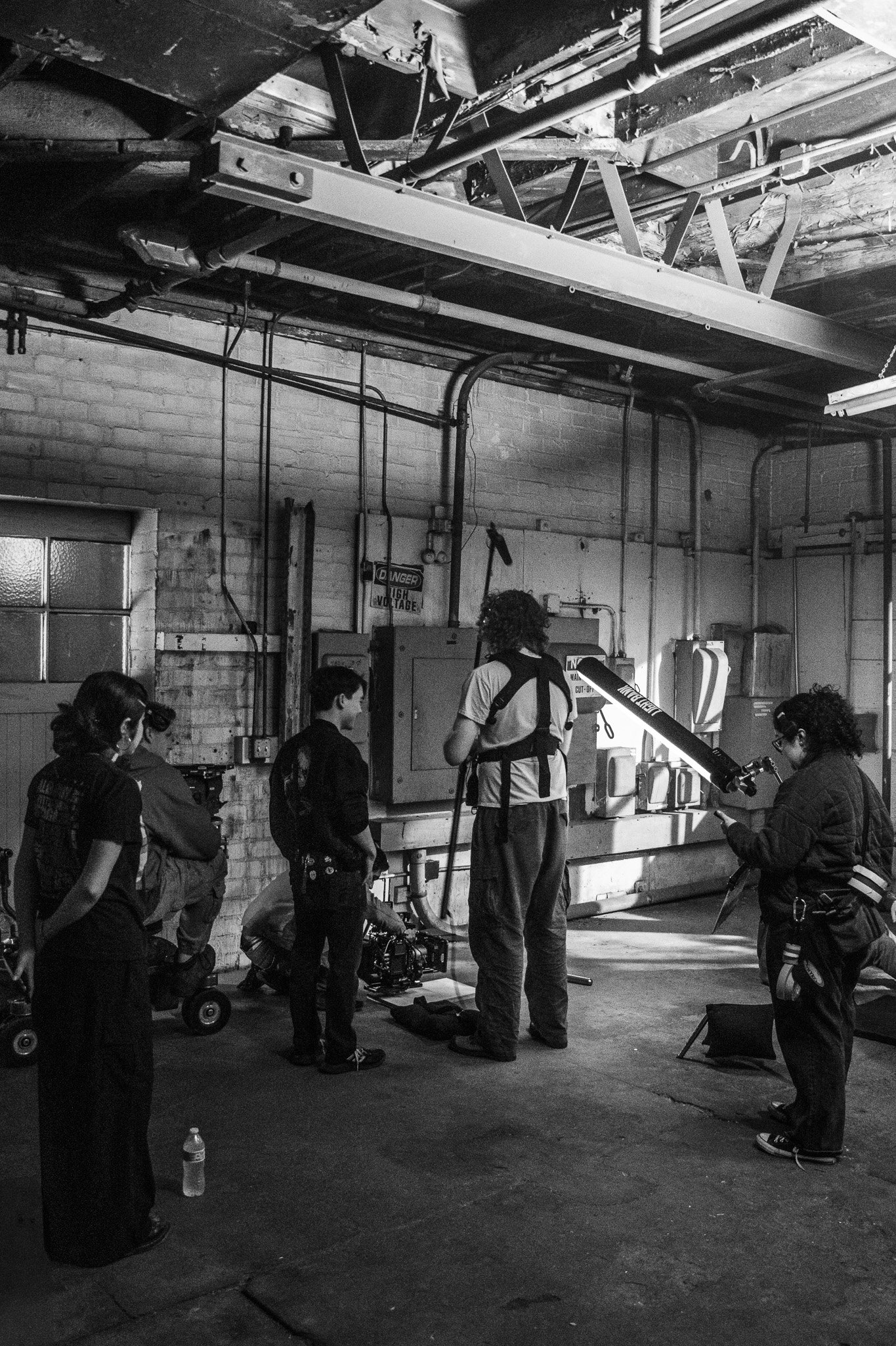 A group of people in a dimly lit industrial or warehouse setting, some wearing headphones, gathered around filming equipment and camera presence. The background shows brick walls, piping, and hanging light fixtures.