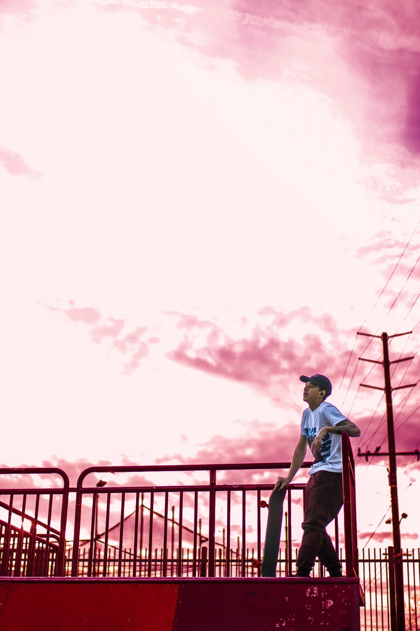 A young man standing on a skateboard ramp under a pink and purple cloudy sky, with power lines and a fence in the background.