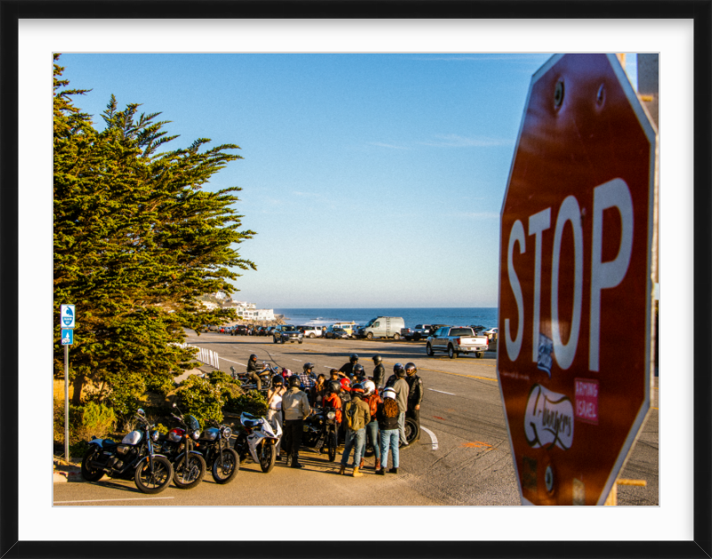 A group of motorcyclists and pedestrians gathered near the curb with motorcycles parked nearby, overlooking a parking lot and the ocean in the background. A stop sign is prominently displayed in the foreground.