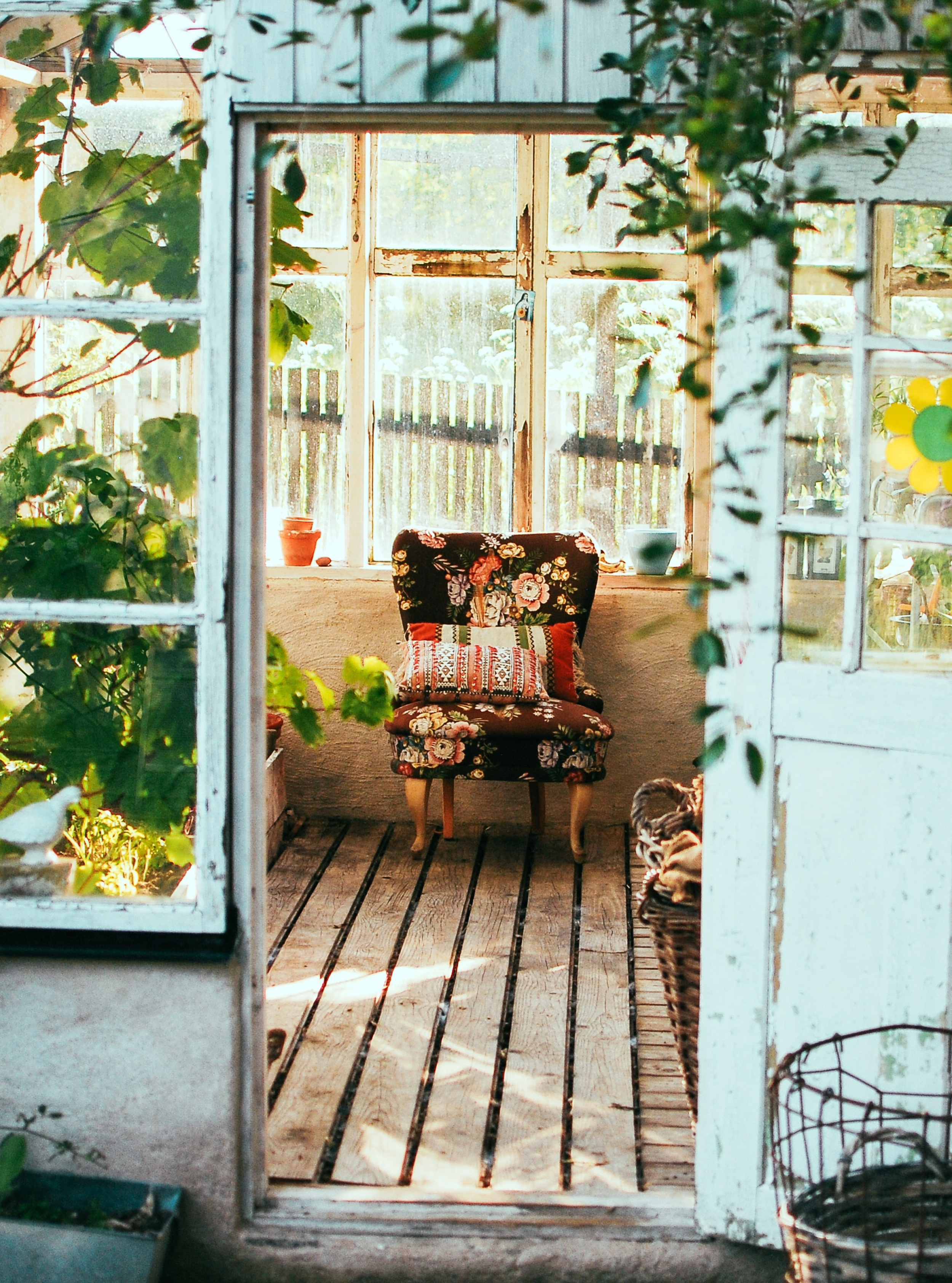 View through an open wooden door into a cozy greenhouse with wooden floors, a floral upholstered chair, potted plants, and sunlight streaming in through large glass windows.