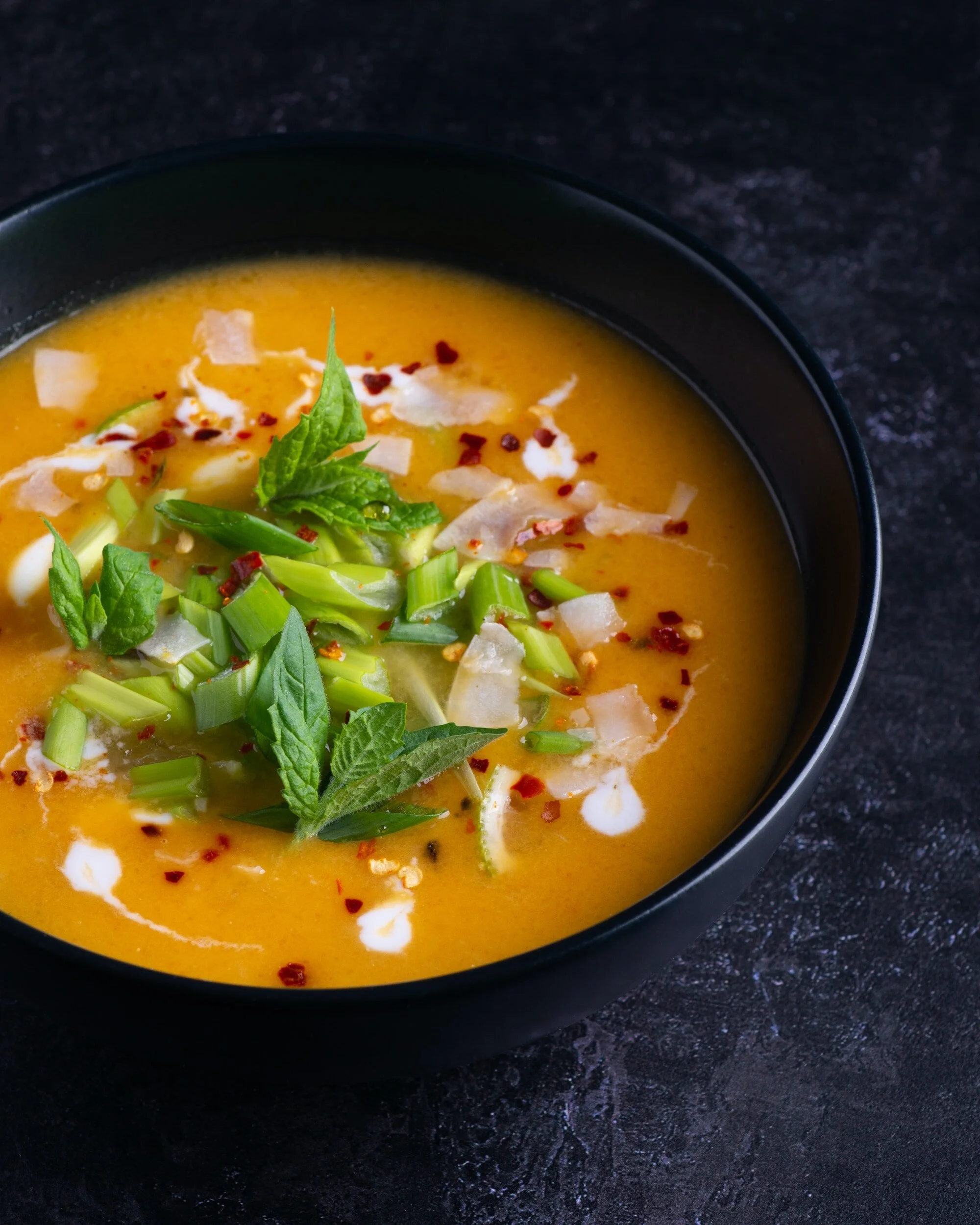 A bowl of orange-colored soup garnished with chopped green onions, fresh herbs, and red chili flakes on a dark surface.