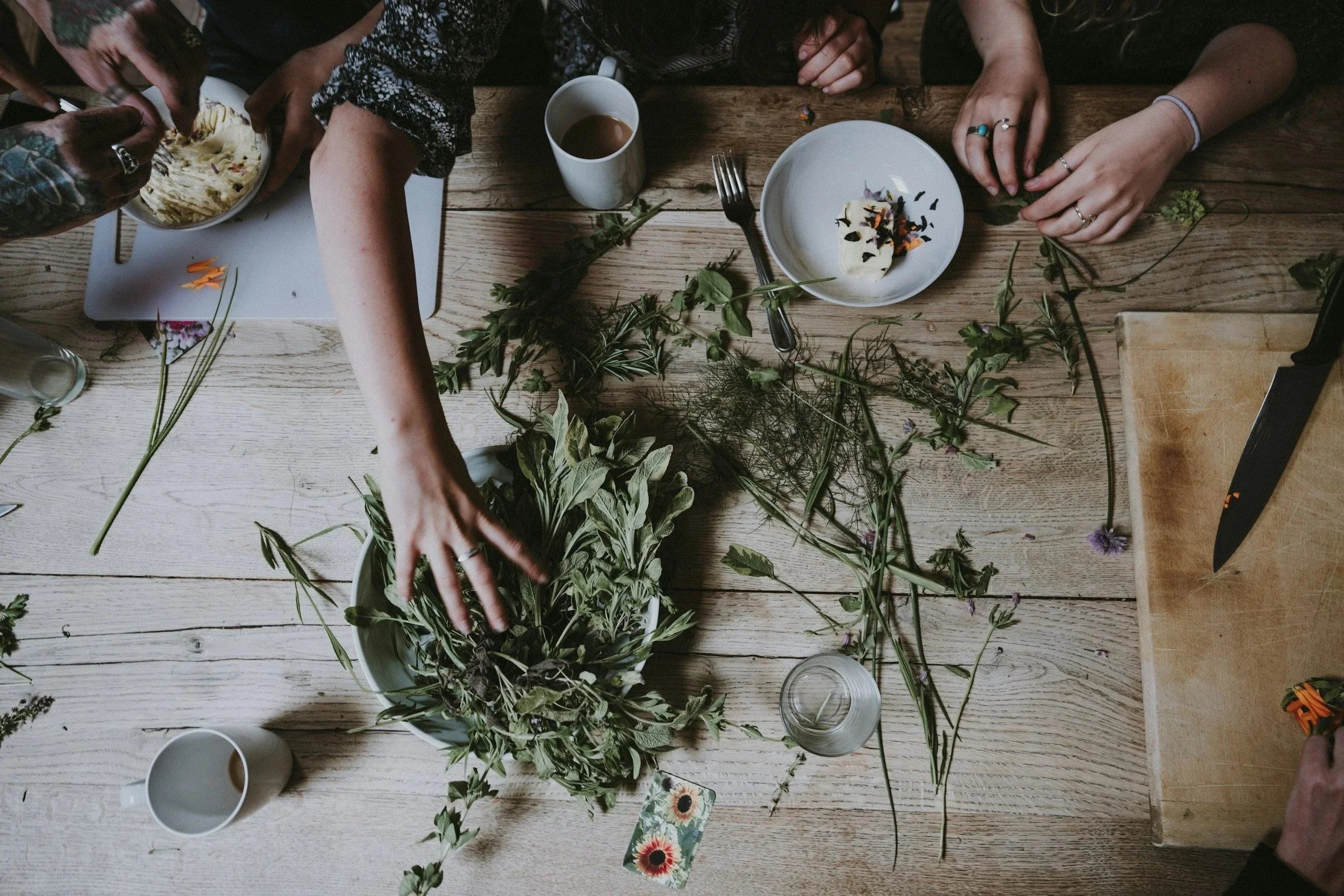 People gathered around a wooden table, arranging and working with various green plants and flowers, with some cups and a bowl of mashed potatoes visible.