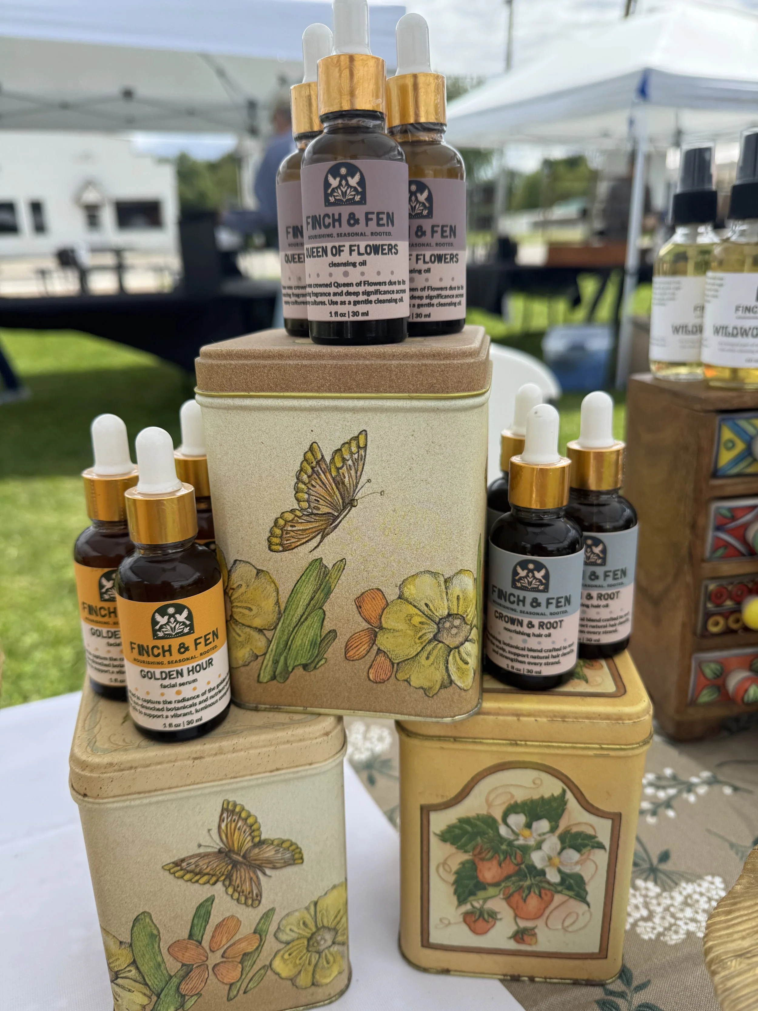 Display of botanical products including facial serum and hair oil bottles with floral and butterfly designs on packaging, set on a wooden stand outdoors.