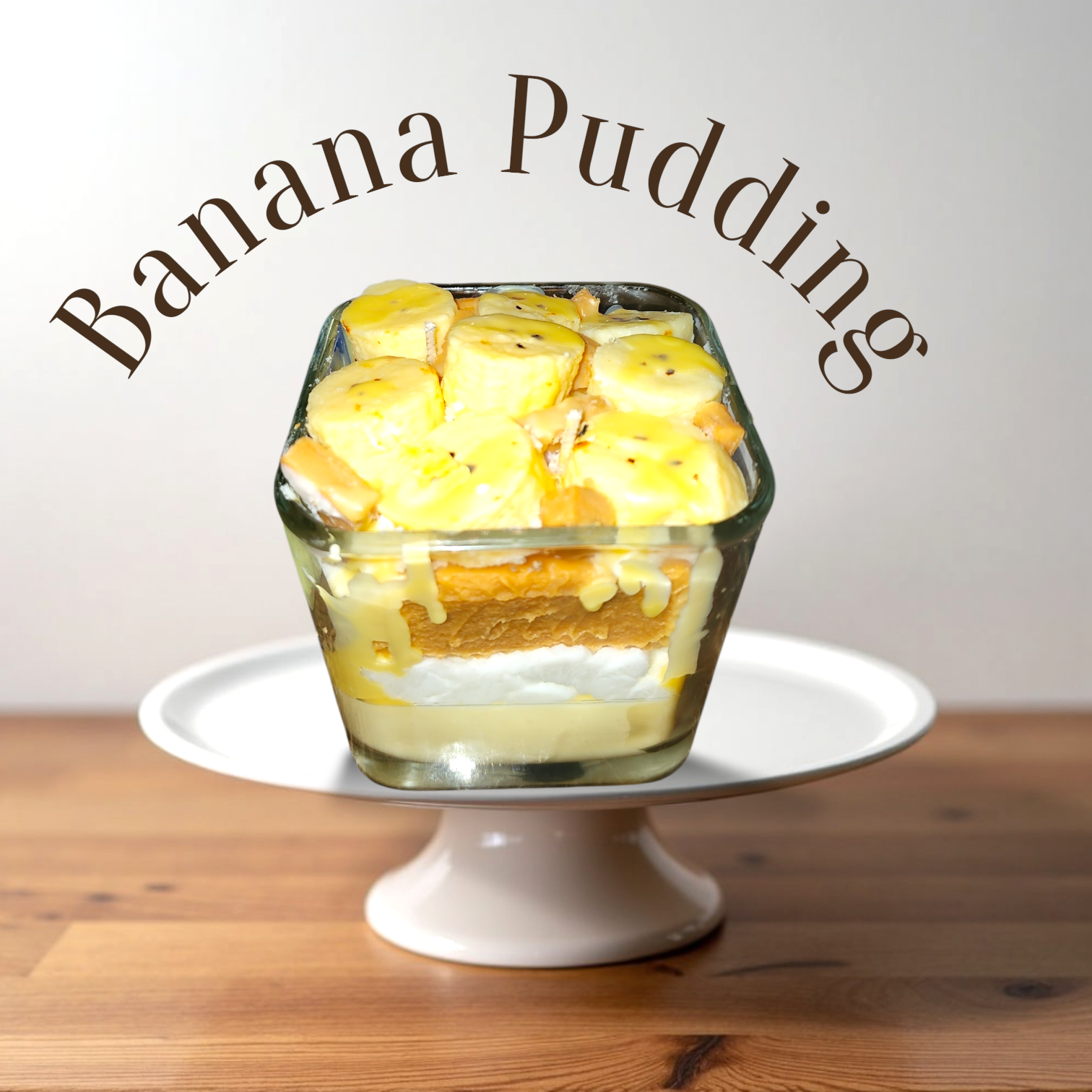 A Dessert Candle banana pudding, layered with sliced bananas, vanilla wafers, pudding, and whipped cream, placed on a white cake stand on a wooden surface with the text "Banana Pudding" above.