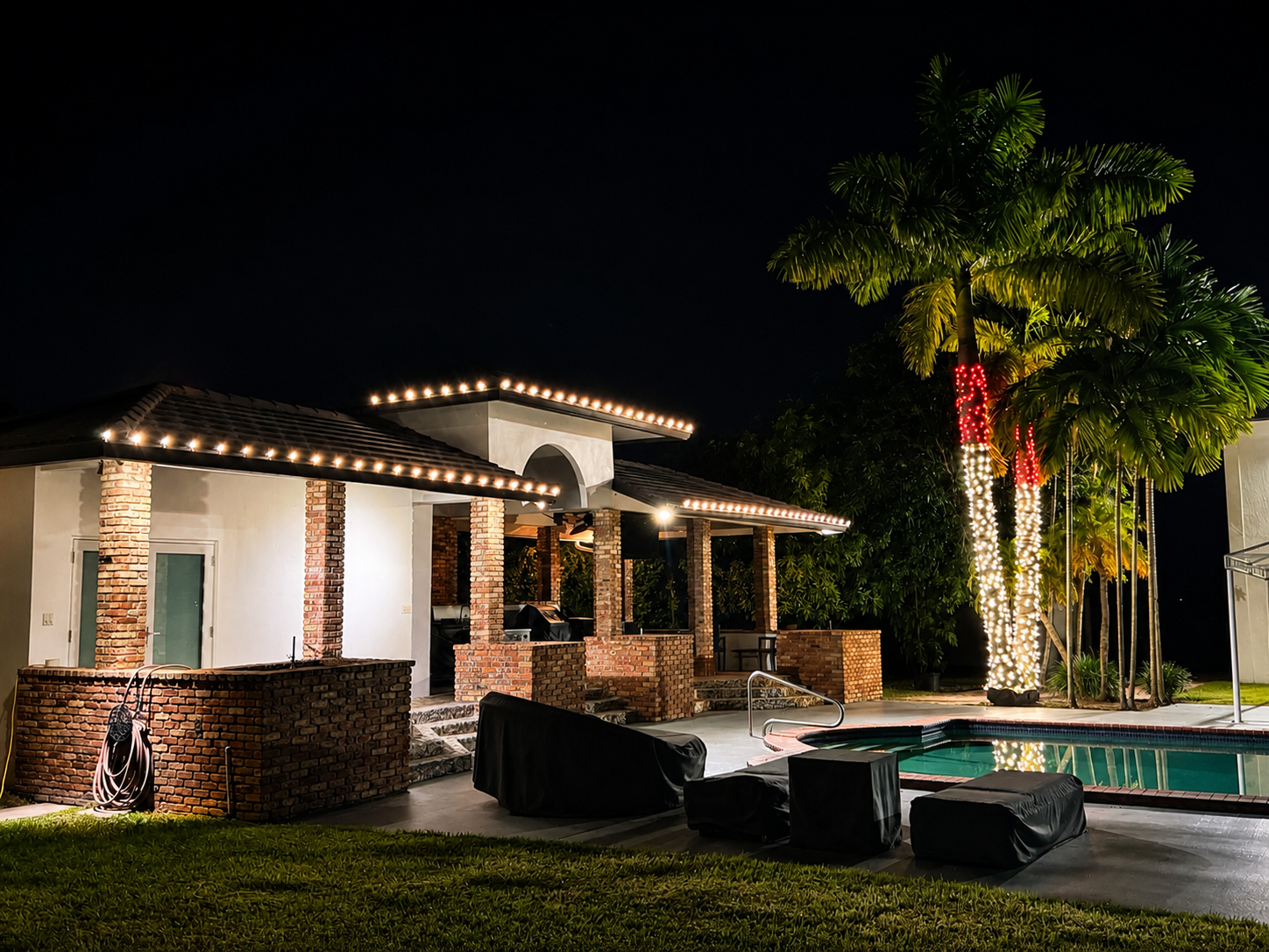 Nighttime view of a backyard with lit pool, covered patio area with string lights, brick columns, tall palm trees wrapped with white and red string lights, and a black covered chair in the foreground.