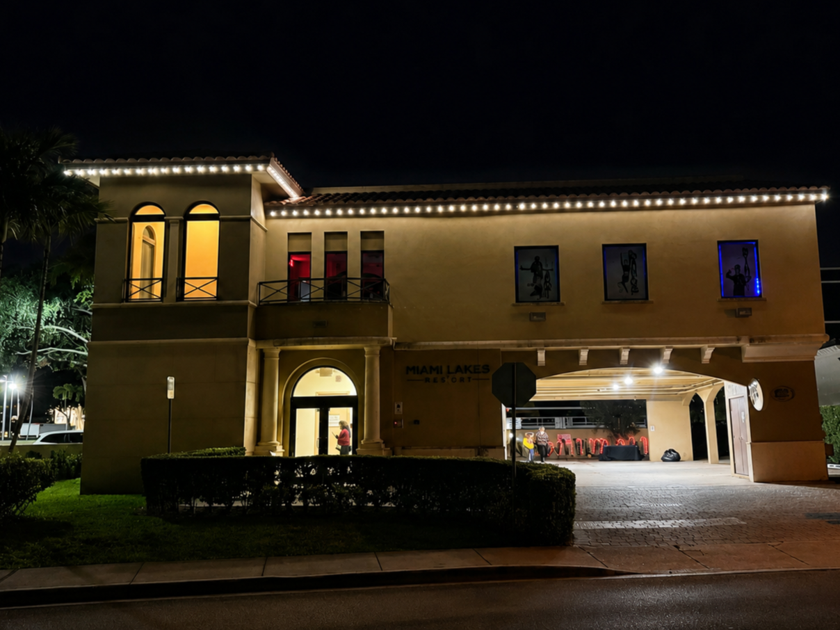 Night view of a two-story building labeled Miami Lakes Resort with decorative lights along the roofline, some windows lit with colorful curtains, and a covered parking area with people and chairs inside.