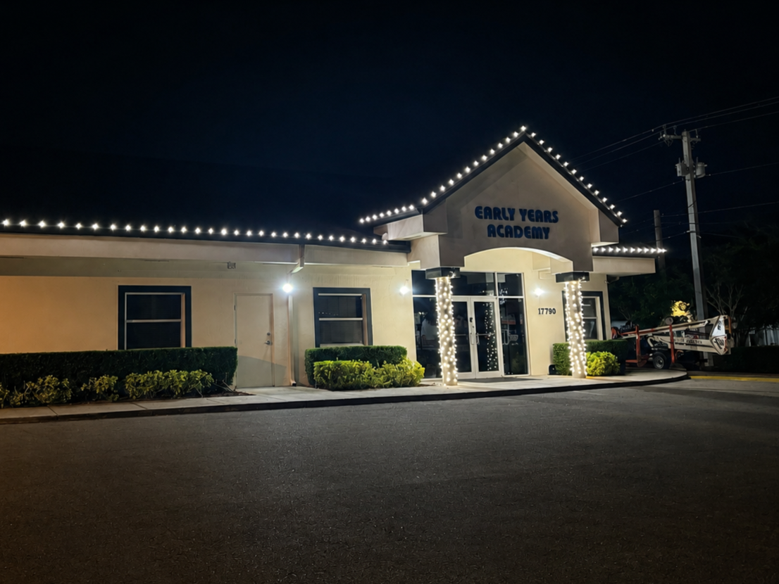 Nighttime view of a single-story building labeled 'Early Years Academy,' decorated with string lights along the roof and around the entrance pillars, with shrubs and a utility crane nearby.