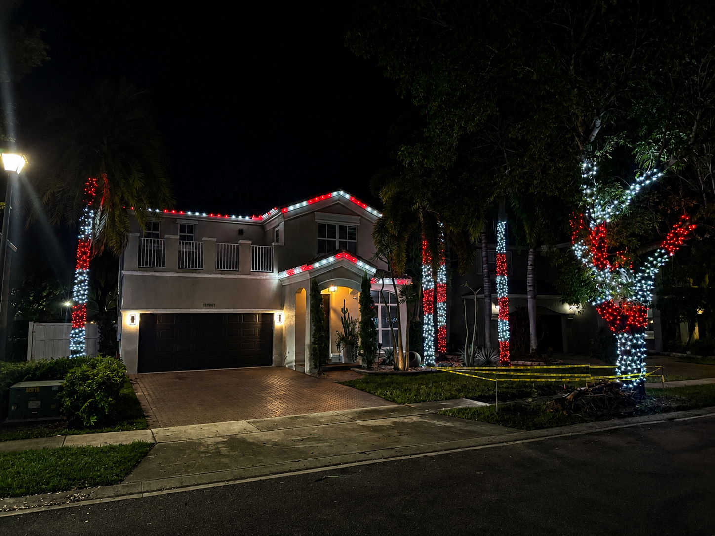 A two-story house decorated with Christmas lights at night. The house has red and white lights along the roof, and trees in the yard are wrapped with similar red and white lights.