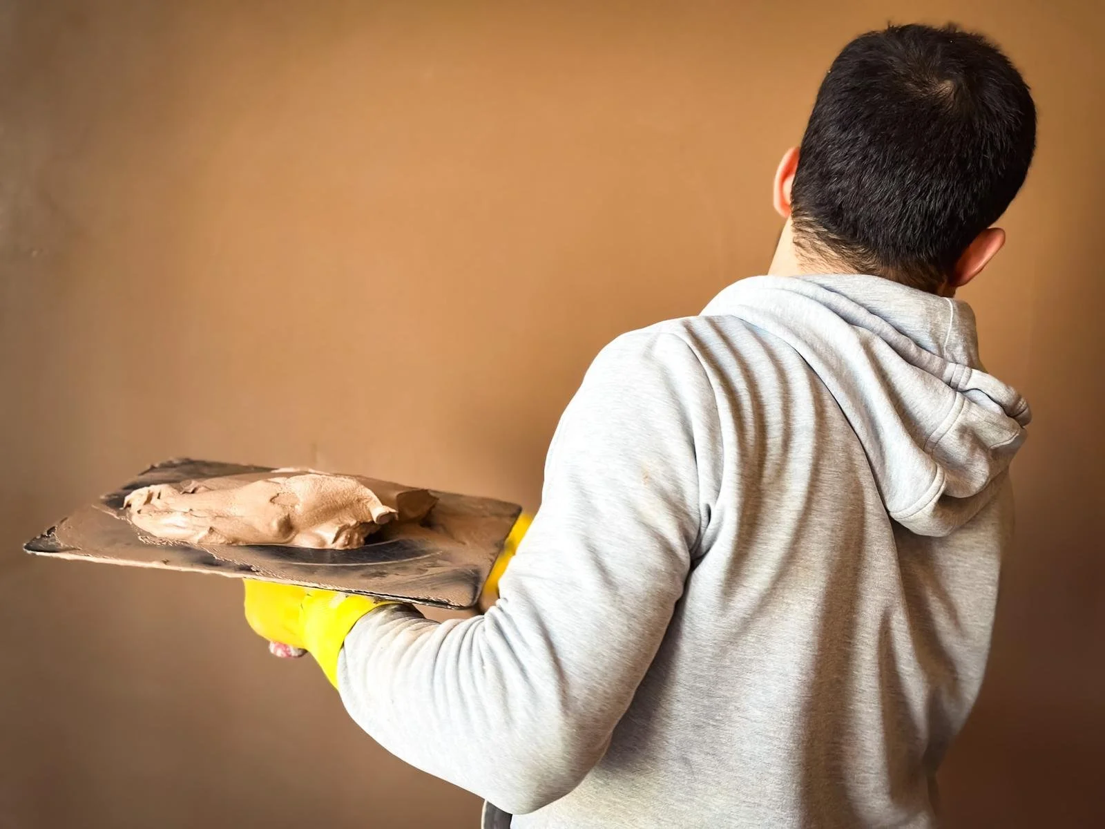 a person viewed from behind, holding a plasterer's hawk in one hand. On the hawk, there's a portion of mixed plaster ready to be applied. The person is wearing a grey hoodie and yellow rubber gloves, which are typically used for protection while work