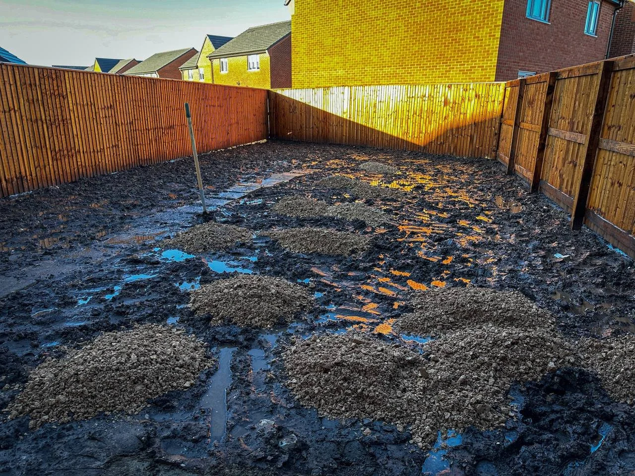 A muddy backyard with a shovel stuck in the ground, surrounded by wooden fences and houses. The scene suggests recent rainfall under a clear, blue sky.