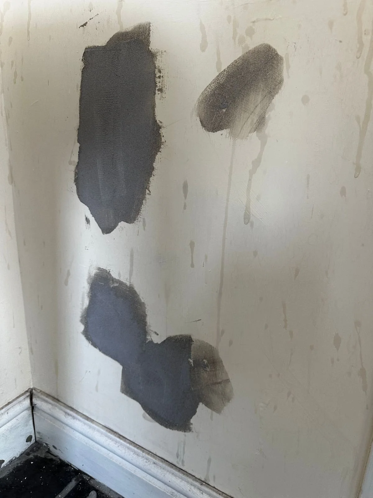 A section of a wall showing plaster patches in progress. Dark grey plaster compound has been applied over larger areas and smaller spots on the wall, indicating the filling of imperfections or damage before the wall is sanded and painted.