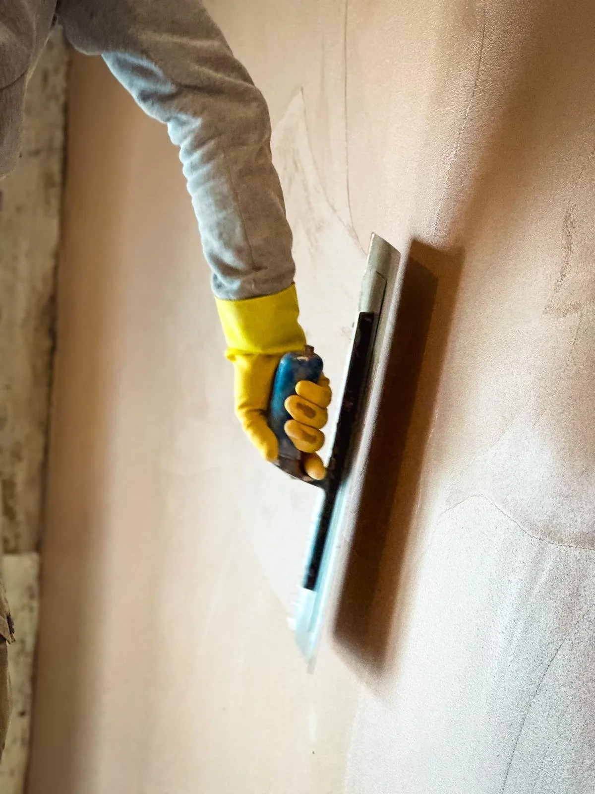 a close-up of a person's arm reaching out to apply plaster to a wall with a blue-handled trowel. The person is wearing a grey long-sleeve top and a yellow rubber glove, suggesting protective wear for construction or renovation work. The angle and foc