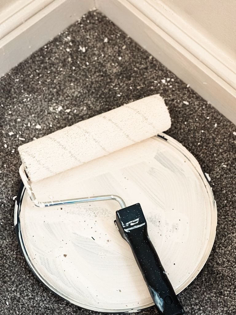 A paint roller covered in white paint rests on an open paint can lid, with splatters of paint visible on the roller, lid, and carpeted floor, indicating a painting task in progress.