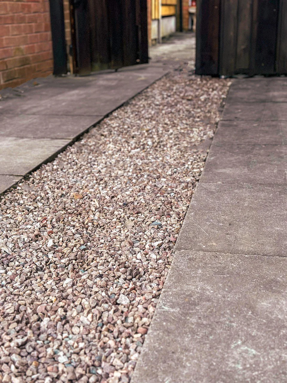 A close-up view of a pathway made of gravel, bordered by larger, weathered paving slabs with a wooden gate in the background
