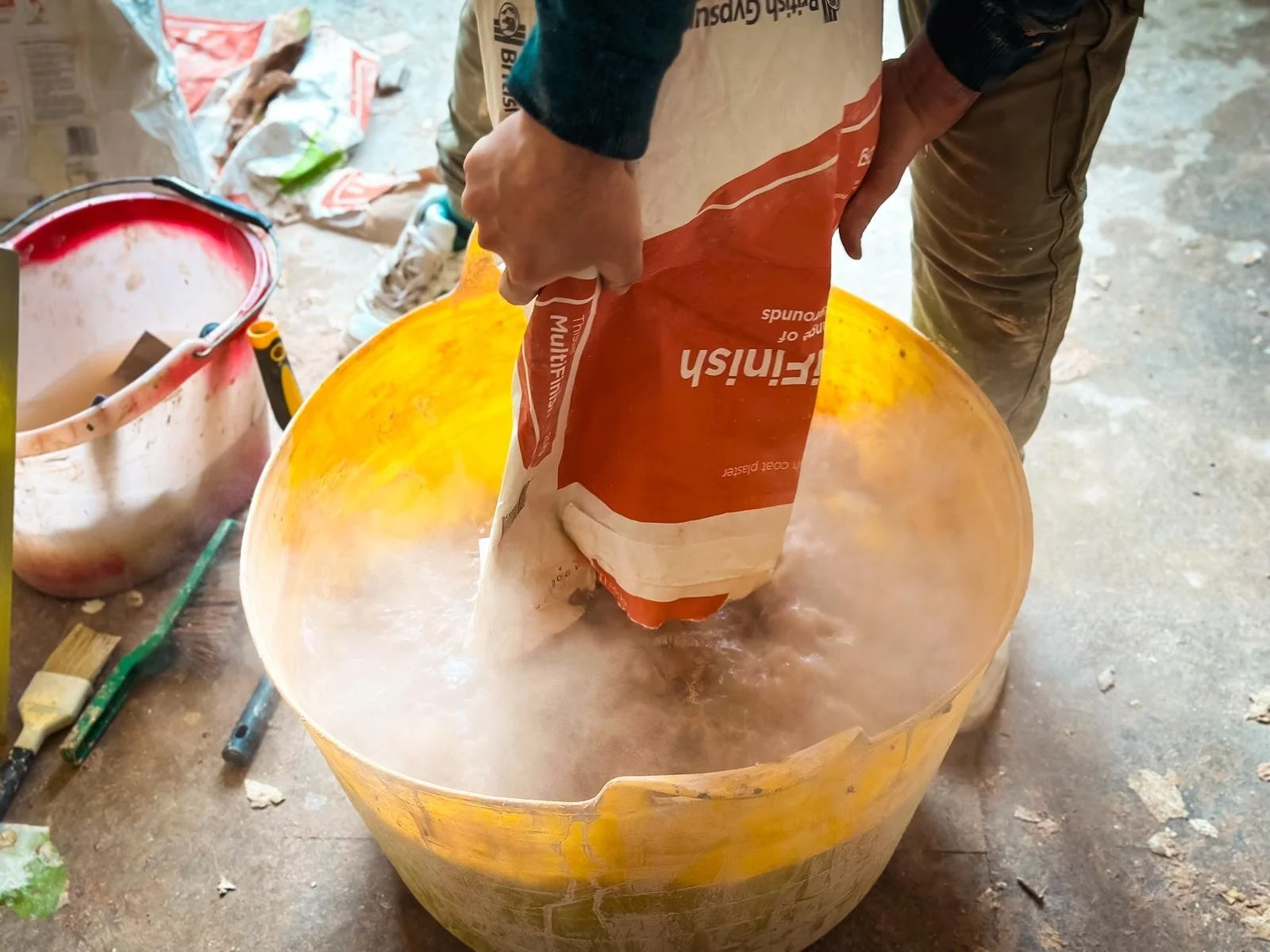 a person pouring dry plaster powder into a large yellow mixing bucket, with a significant amount of dust being generated as the powder is emptied from the bag. The person is wearing casual clothing, suggesting DIY work rather
