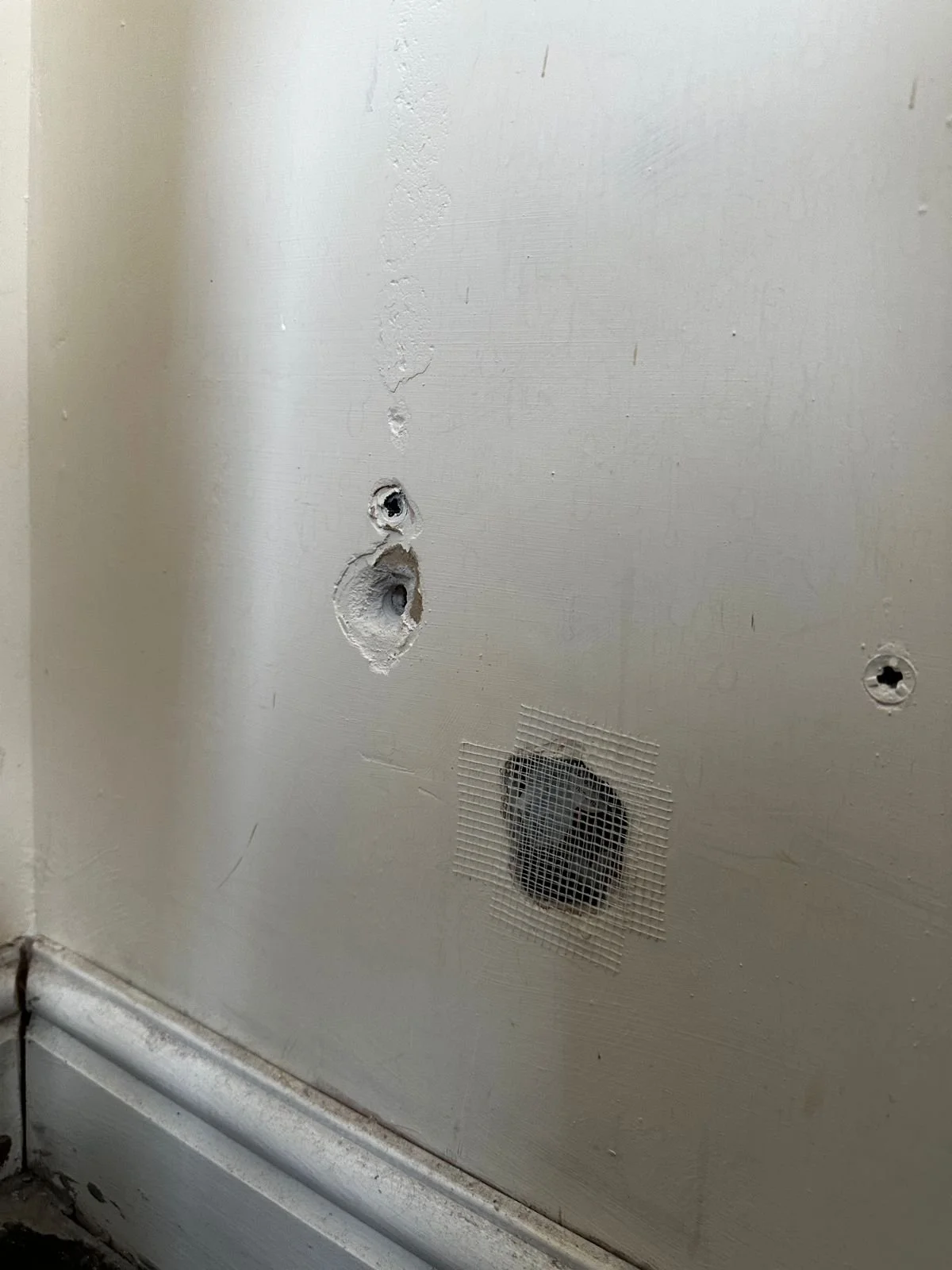 A close-up of a white plasterboard wall with some repair work visible. Two holes have been patched with plaster, and a square section is covered with a mesh tape, commonly used to reinforce the plaster repair and prevent cracking.