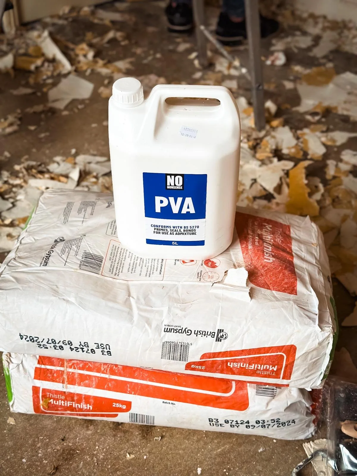 a 5-liter container of "NO NONSENSE PVA" adhesive placed on top of a wrapped 25kg bag of Thistle MultiFinish plaster. The scene is a construction or renovation site, as indicated by the scattered debris on the floor and the legs of a stepladder in th