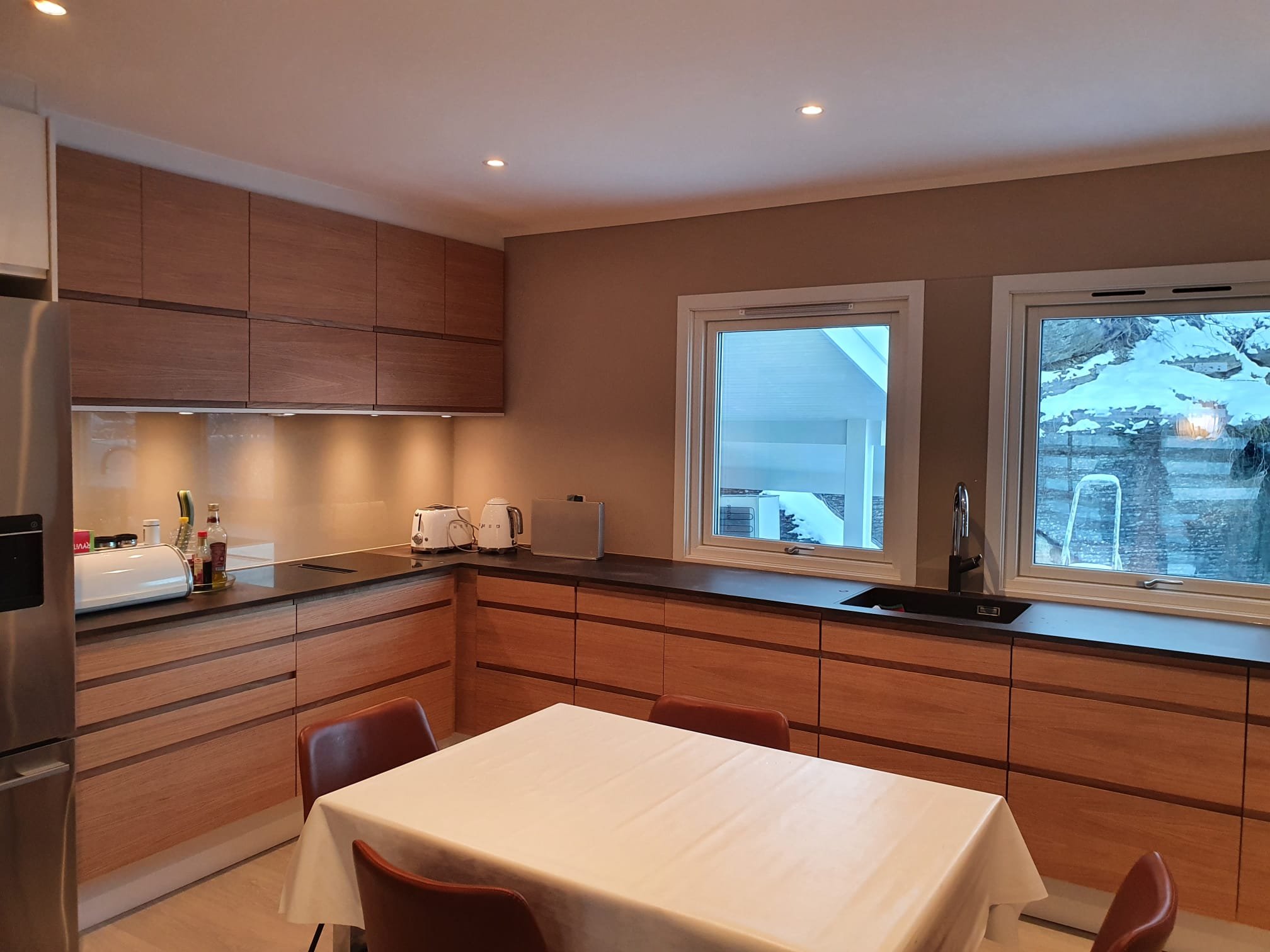 A modern kitchen with wooden cabinets, built-in appliances, and a dining table, with a view of snow outside through the windows.