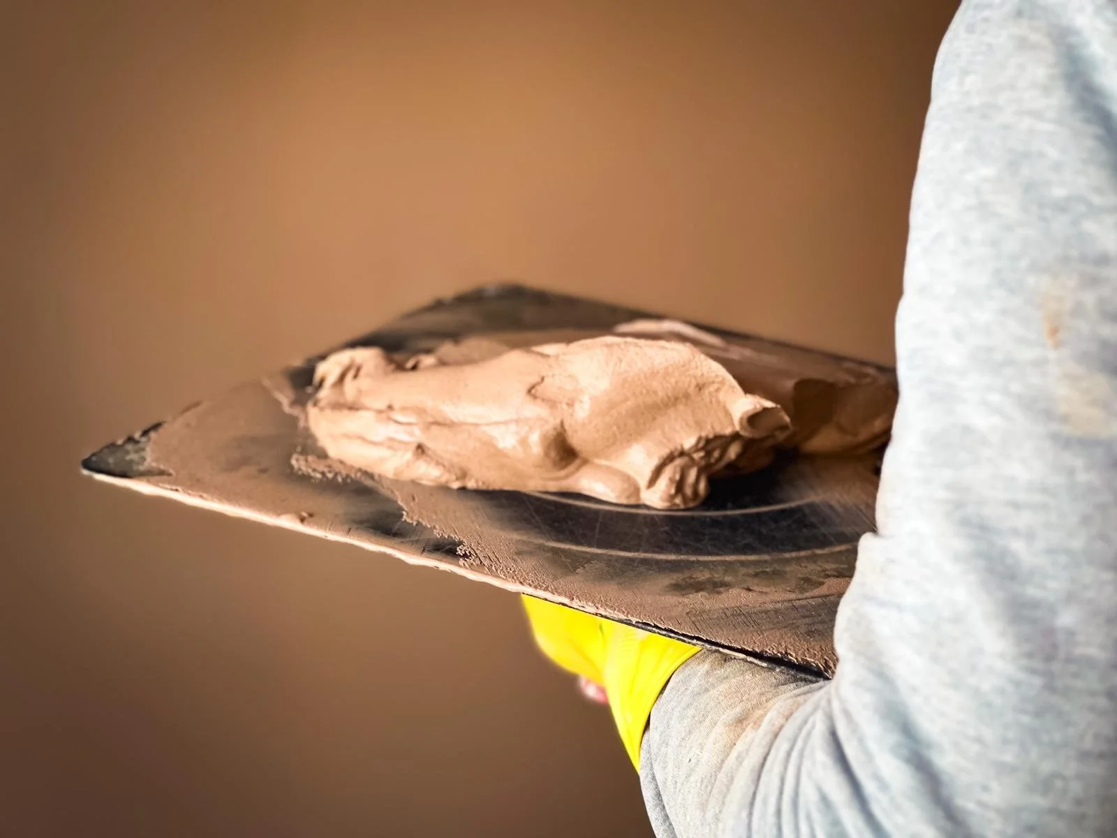 a close-up of a person holding a plasterer's hawk with a dollop of freshly mixed plaster on it. The hawk appears well-used, which is common for a tool that's an essential part of a plasterer's kit. The person is wearing a grey long-sleeve shirt and