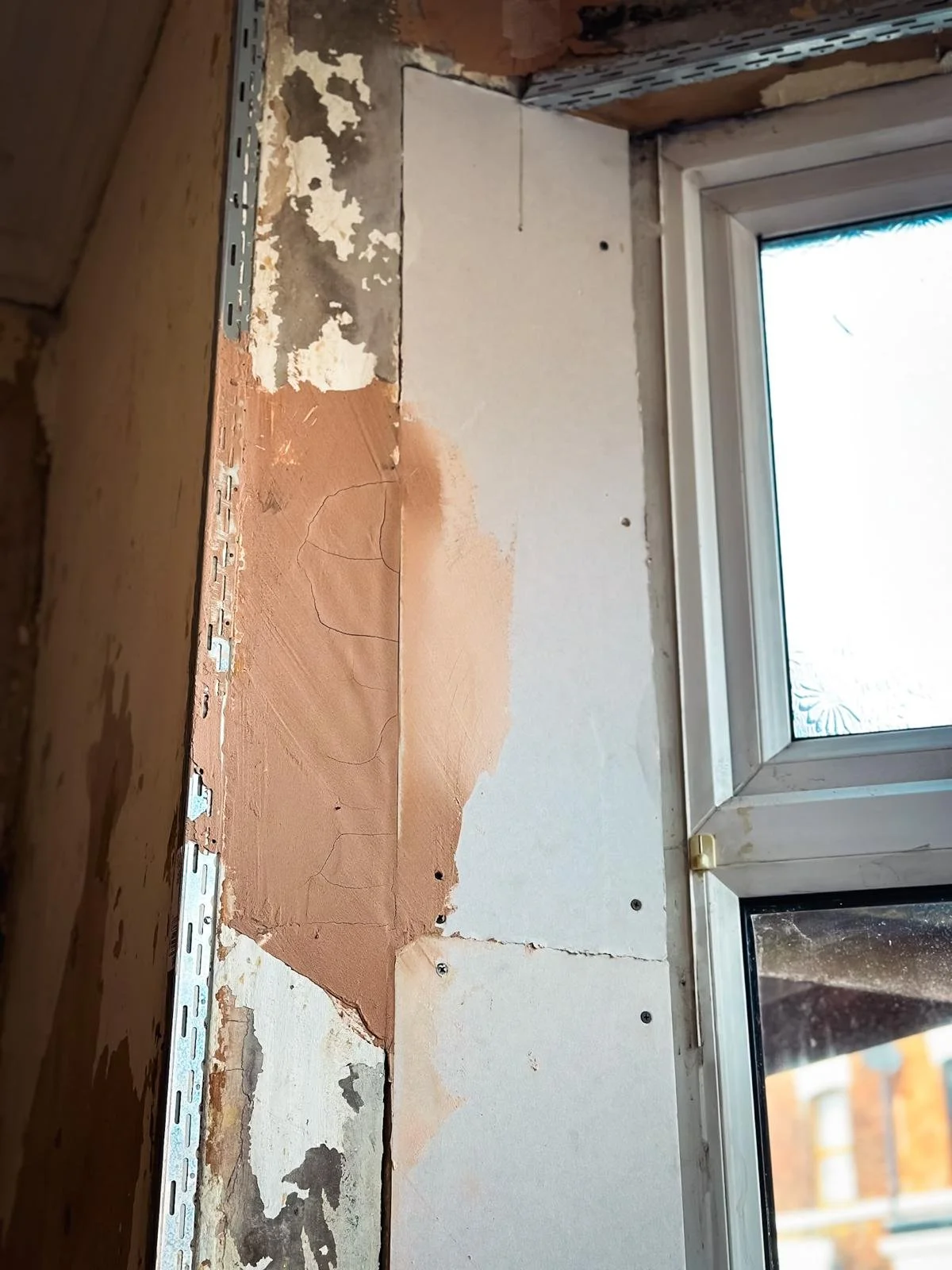 a close-up view of a window frame during what appears to be renovation work. The wall surrounding the window has been partially stripped, revealing layers of old paint and patches of bare plaster. To the left, there's a metal corner