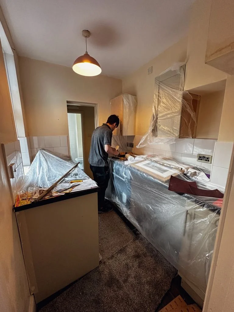 A person in a kitchen focusing on a task at hand, possibly repairing a window or working on a DIY project. The kitchen is covered with protective plastic sheeting to shield appliances and surfaces from dust and debris
