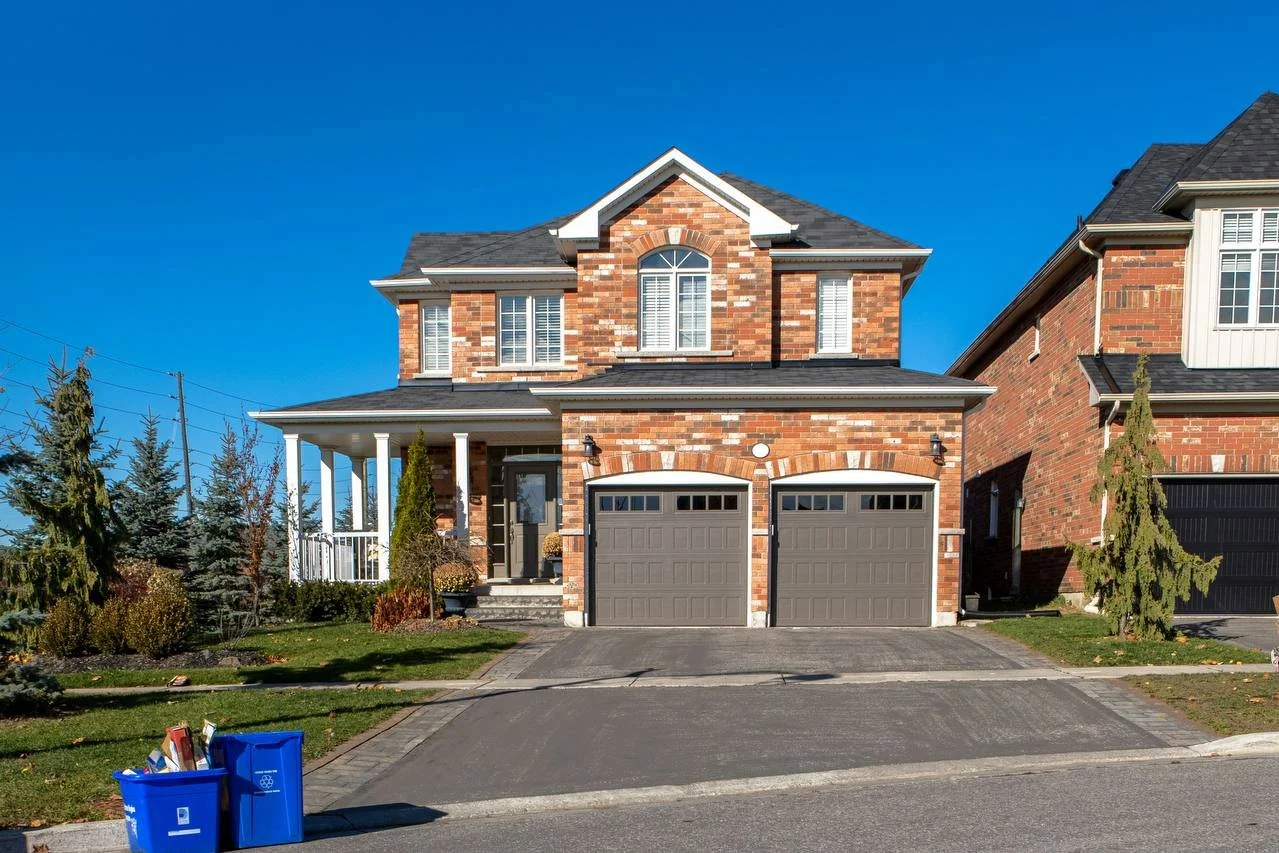 two-story brick house with a well-maintained front yard and two recycling bins on the driveway. The house features two garage doors, a covered porch, and several windows.