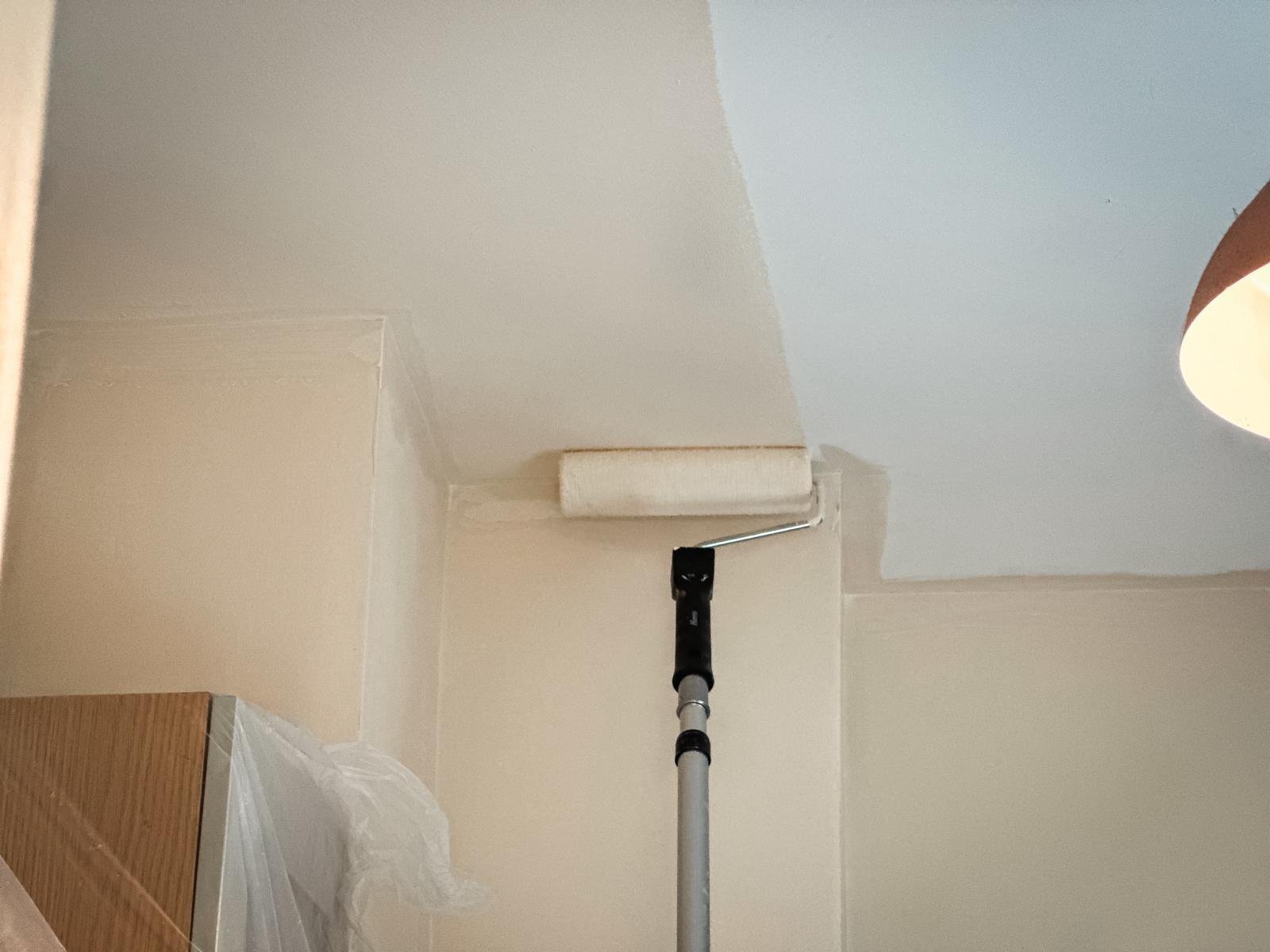 White wall and ceiling being painted with a creamy paint using a paint roll