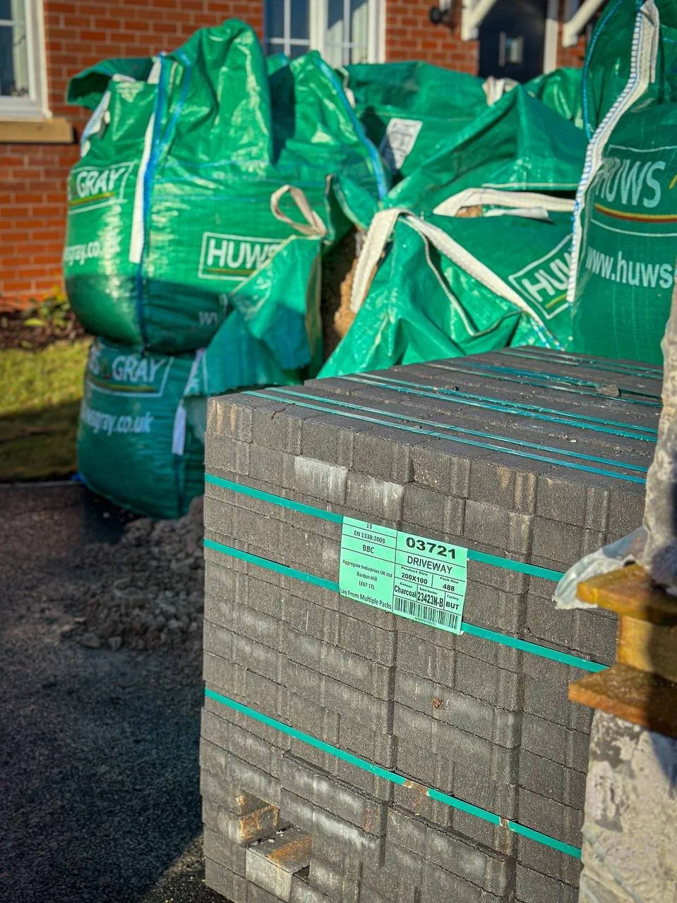 A stack of driveway paver stones with a label, in front of large green waste bags labeled 'Huws Gray', likely at a construction or landscaping site.