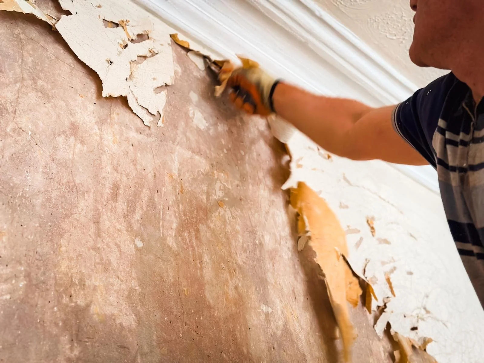 A person is scraping off old, peeling wallpaper from a wall. The wall underneath has a patchy appearance with remnants of glue or wallpaper, and the person is using a tool in their right hand to remove the wallpaper. The scene suggests a home renovat