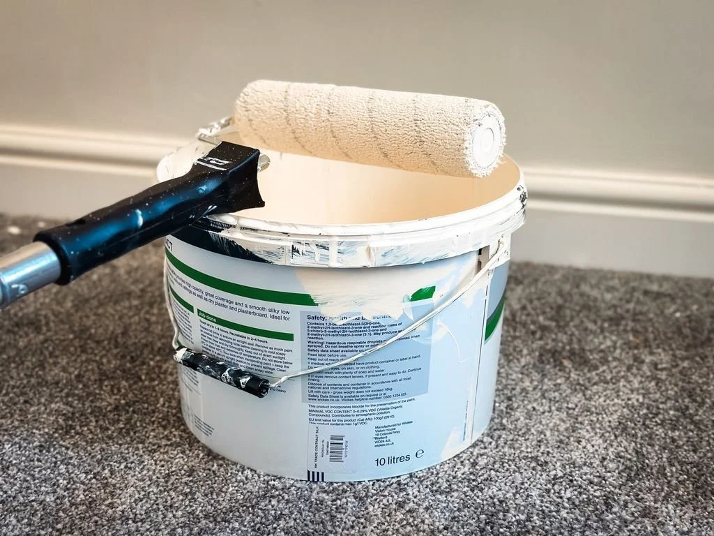 a 10-litre bucket of paint with a paint roller resting on top. The roller has a thick nap cover, which is commonly used for painting textured surfaces or for applying paint over rough plaster. The bucket appears to have been used, with paint residue