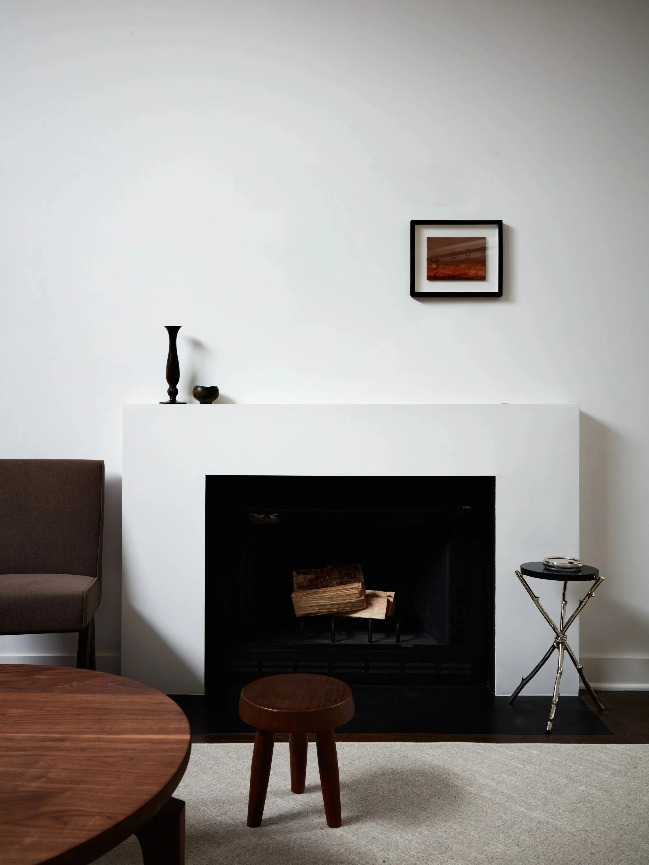 Living room with a fireplace, a piece of wall art, and decorative items including a black vase and a small bowl on the mantle, a brown side table and stool, and a metal and glass side table.