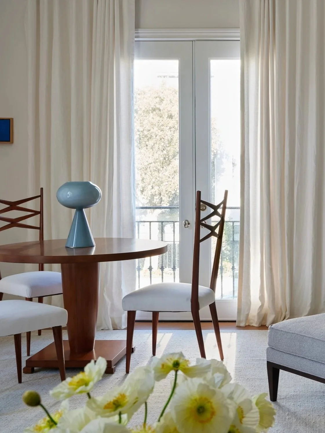 Interior view of a dining area with a round wooden table, white cushioned chairs, a blue lamp on the table, cream curtains, and a glass door leading to a balcony with trees outside.