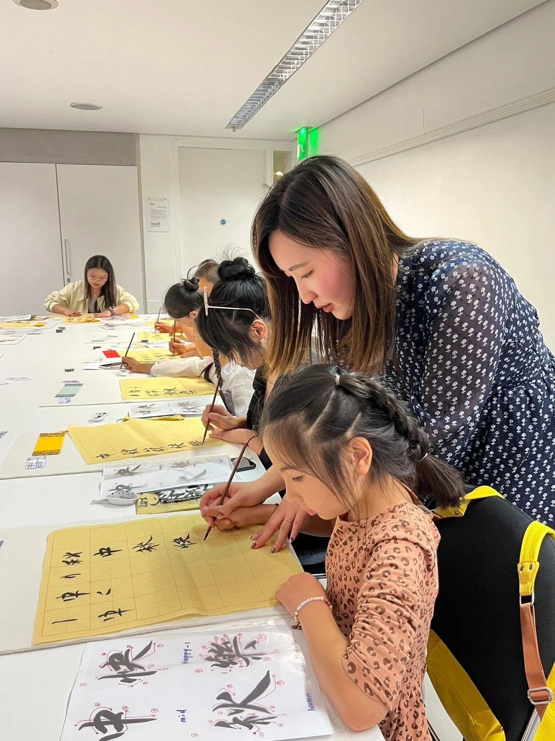 A group of people practicing Chinese calligraphy at a table, guided by an instructor, in a classroom setting. Workshop teacher: Yi Ling Lai, the founder of lingsspace, Location: The British Museum, London