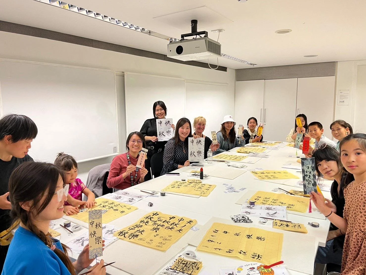 A group of people seated around a table practicing calligraphy, displaying their work and holding up papers with Chinese characters. Workshop teacher: Yi Ling Lai, the founder of lingsspace, Location: The British Museum, London