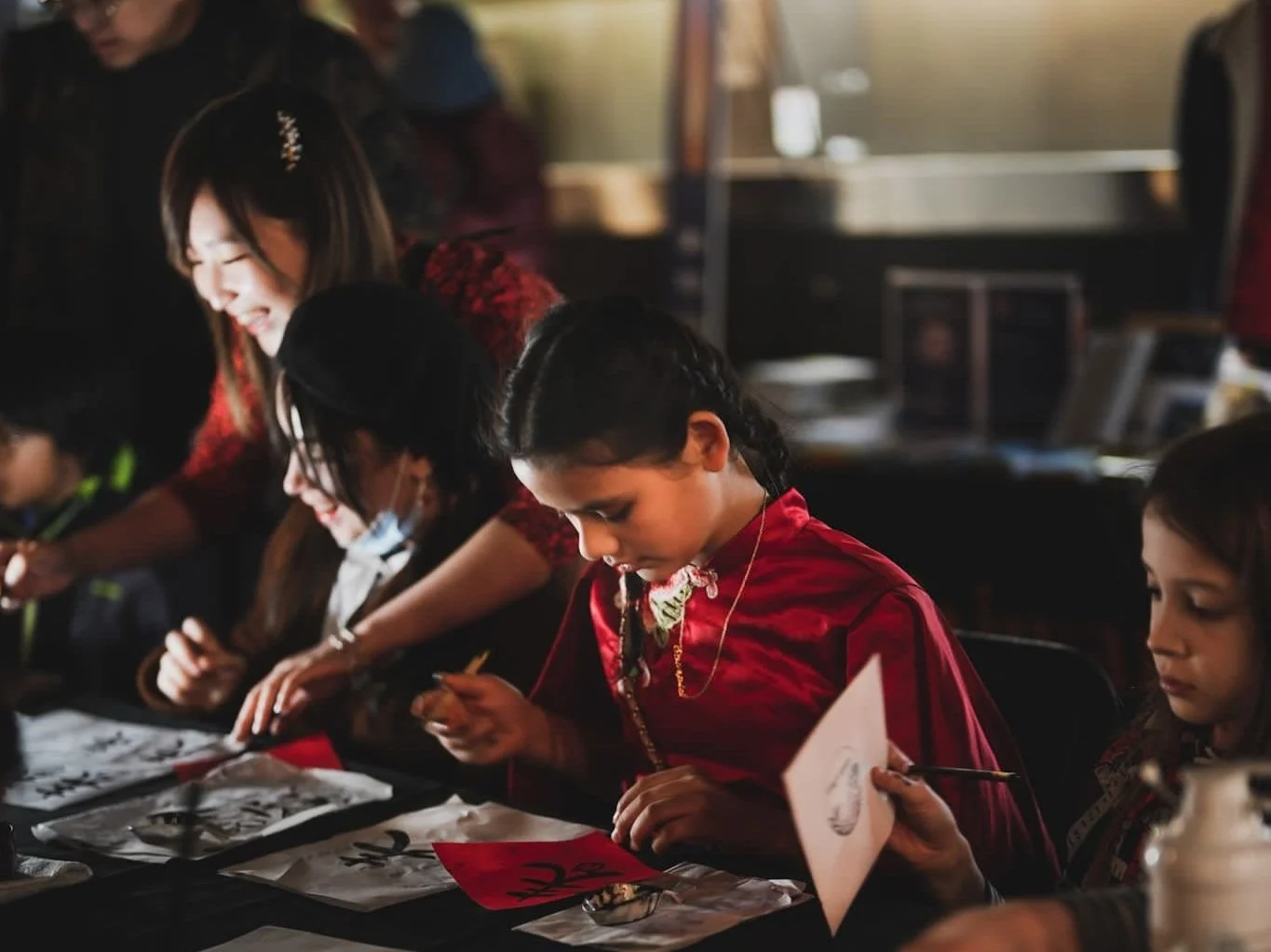 Children engaged in calligraphy activity, wearing red traditional clothing, with brushes and paper, guided by an adult woman. Indoor setting. Workshop teacher: Yi Ling Lai, the founder of lingsspace