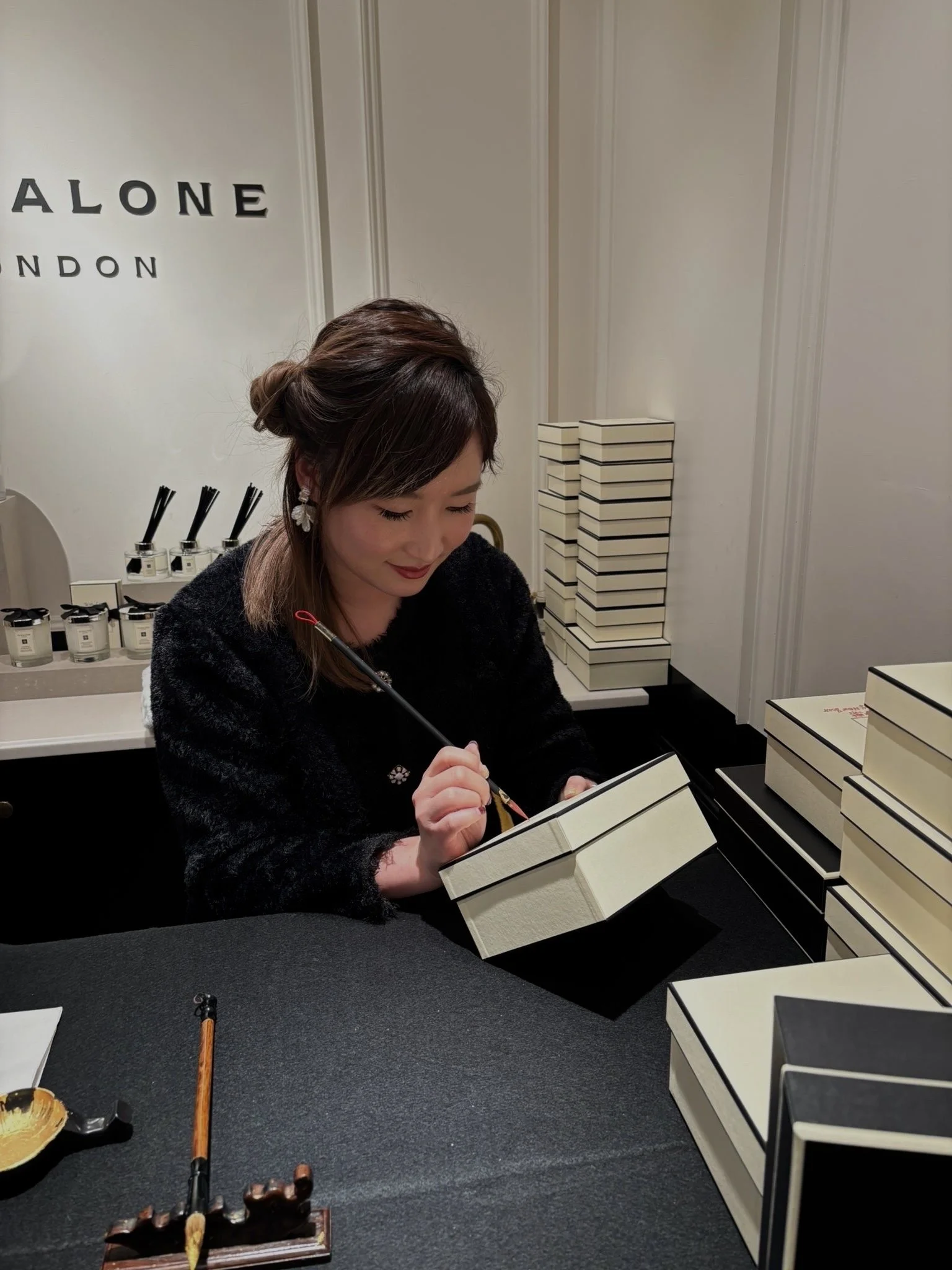 A person writing on a box in a Jo Malone store with black and white packaging stacked on a table. Artist: Yi Ling Lai, the founder of lingsspace