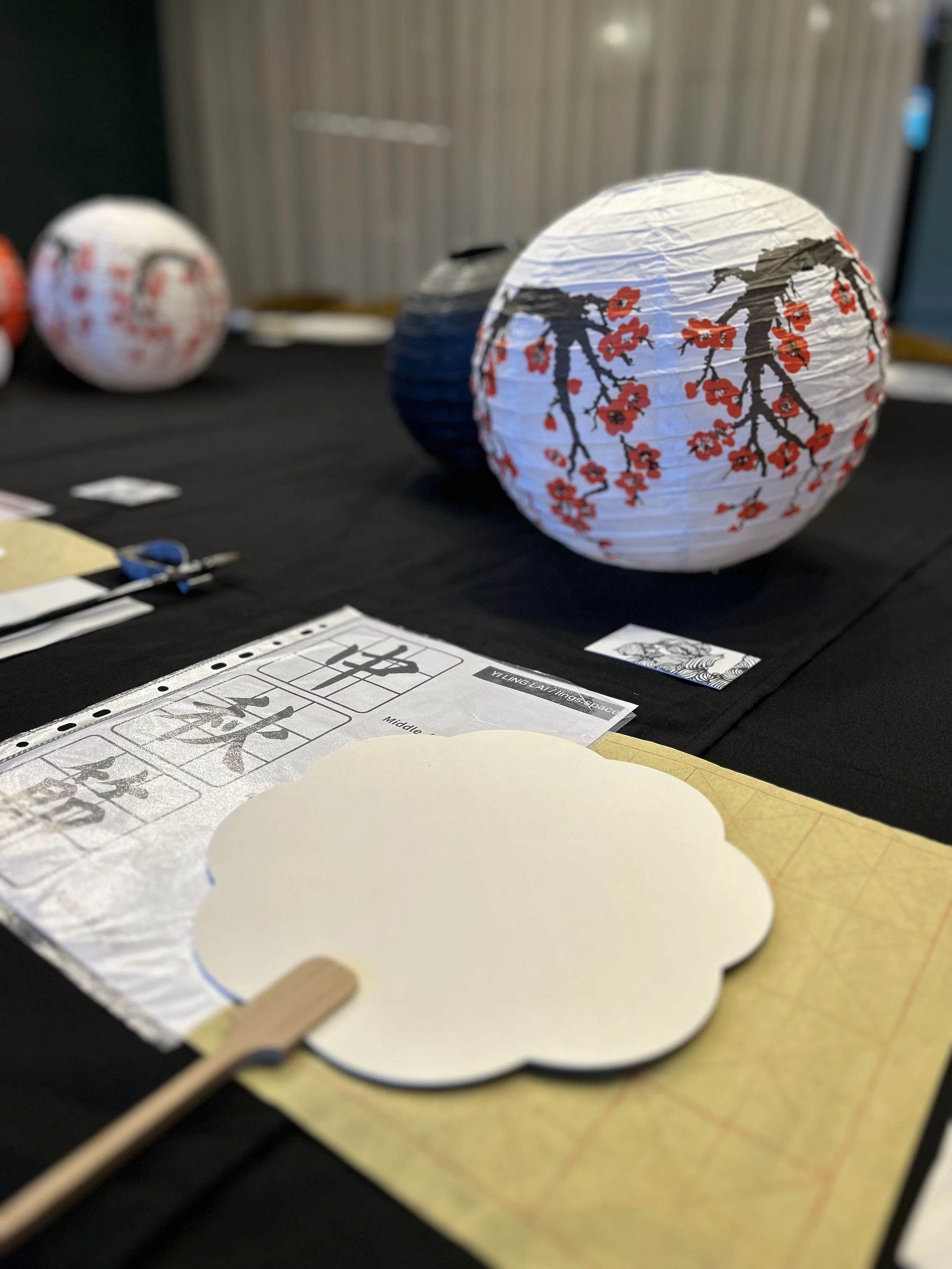 Company workshop: A close-up of a Chinese-style table setup featuring a white paper lantern decorated with red cherry blossoms and black branches, set on a black surface alongside a blank fan template and practice calligraphy sheets.