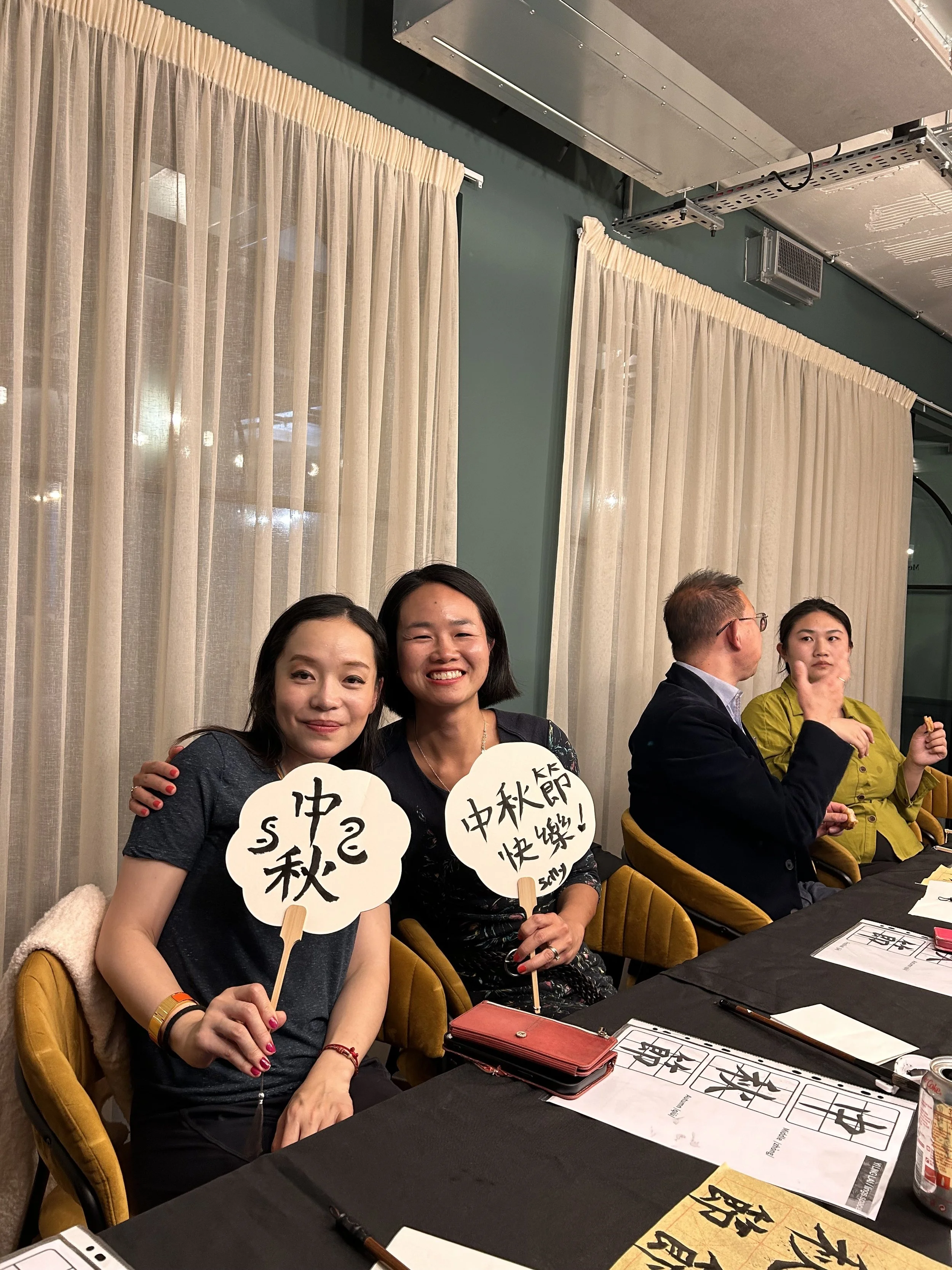 Company workshop: Two women sitting at a table holding signs with Chinese characters, surrounded by others at a social event indoors. workshop teacher: Yi Ling Lai, the founder of lingsspace